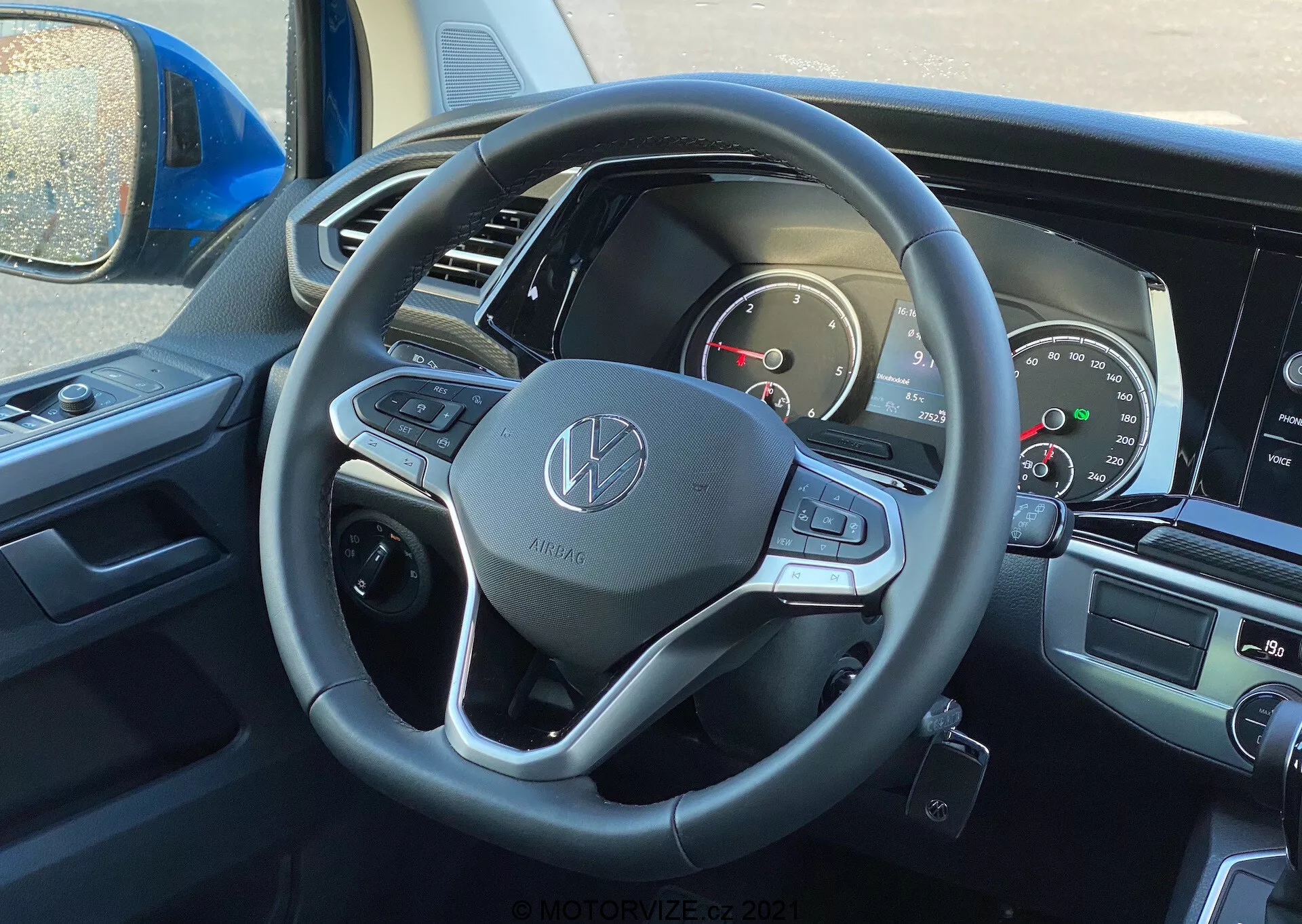 Pogled na notranjost VW Nutzfahrzeuge Transporter 2019 (T6-1 Multivan) iz perspektive voznika, s poudarkom na volanu in armaturni plošči. Volan prikazuje emblema Volkswagen in je opremljen s usnjenim ovitkom, večfunkcijskimi gumbi in obvolanskimi ročicami za prestavljanje. Delno viden instrumentni panel vključuje analogne števce in digitalni zaslon. Centralni informacijsko-zabavni zaslon in krmilniki klimatske naprave so rahlo vidni, kar nakazuje na moderne multimedijske in udobnostne funkcije.
