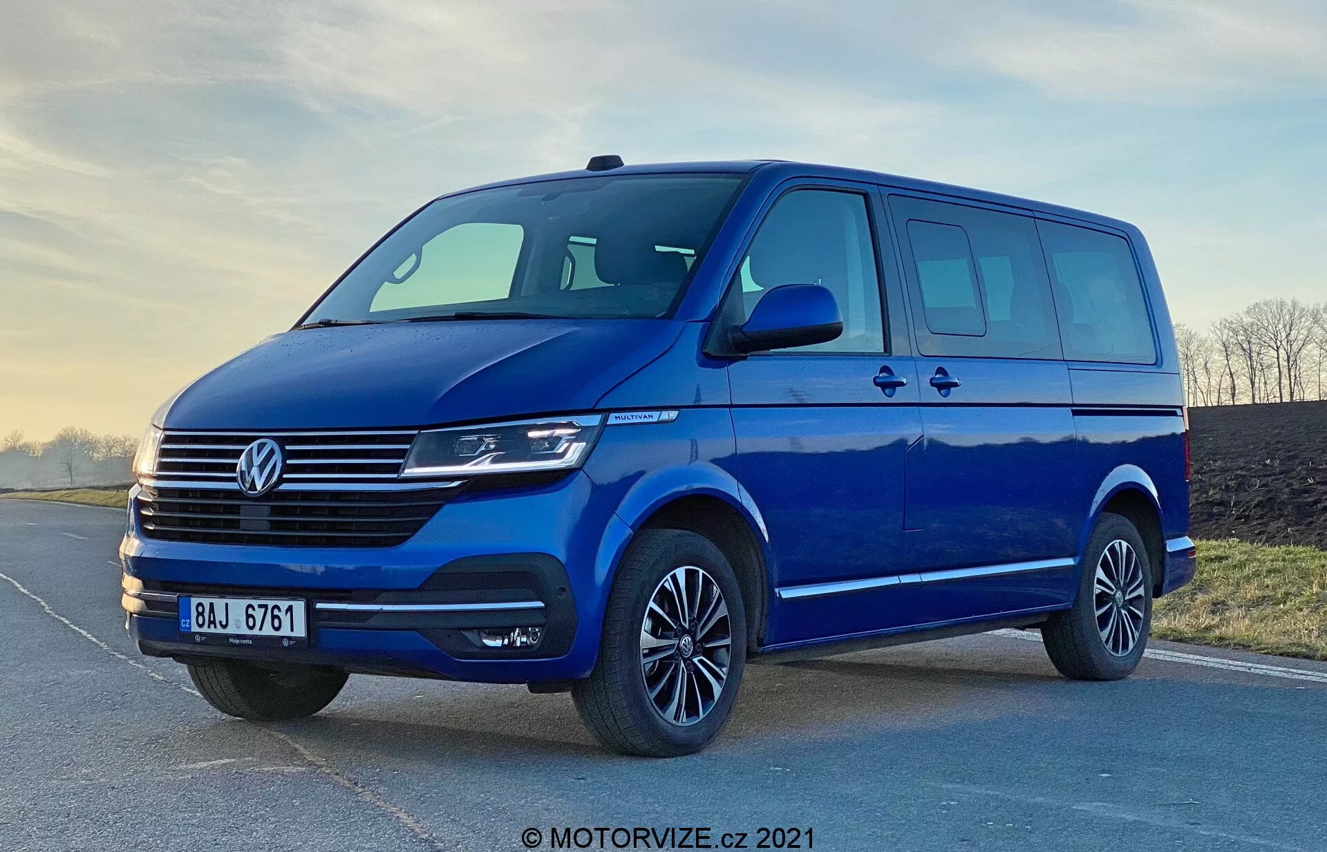 Slika modrega Volkswagen Transporter T6.1 Multivan iz leta 2019, posneta iz nizkega kota, da poudari njegovo postavo in velikost. Sprednji tričetrtinski pogled prikazuje sprednji in stranski dizajn vozila, vključno z značilno rešetko Volkswagen z vodoravnimi črtami, osrednjim emblemom, sodobnimi žarometi, ki verjetno vključujejo tehnologijo LED ali Xenon, in elegantnim sprednjim odbijačem s širokim dovodom zraka. Aerodinamična stranska ogledala z vgrajenimi smerniki, veliko okenskega prostora z drsnimi vrati na strani, aluminijasta platišča z nizkoprofilnimi pnevmatikami in mehka osvetlitev poudarjajo praktičnost in estetsko privlačnost kombija. Vodni žig 'MOTORVIZE.cz 2021' kaže, da je slika iz leta 2021.