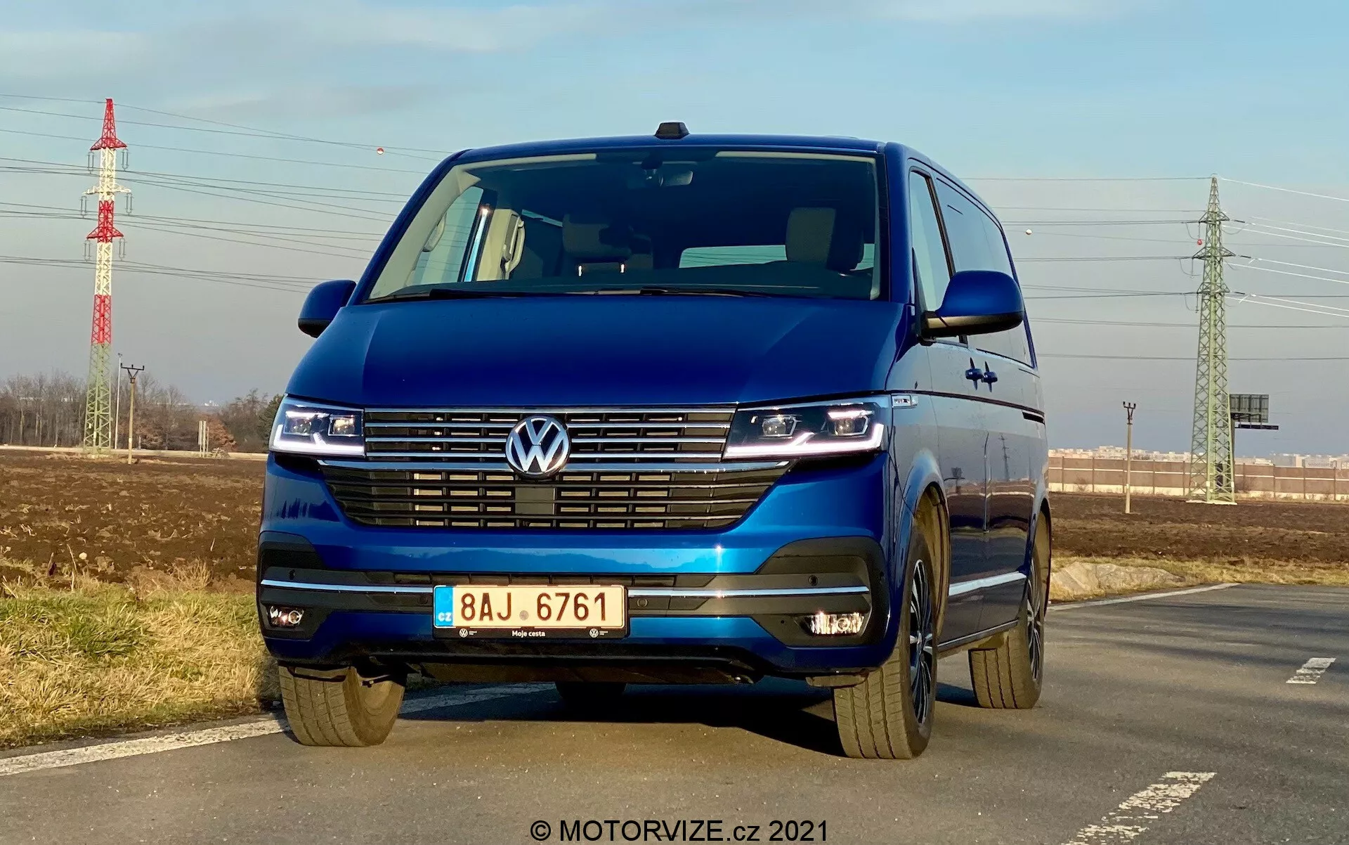 Modri ​​Volkswagen T6-1 Multivan 2019 ujet iz nizkega sprednjega kota, ki poudarja masko, LED žaromete z dnevnimi lučmi, Volkswagenov emblem, široko masko, dvobarvni odbijač in aluminijasta platišča; parkiran ob cesti, z jasnim nebom, oddaljeno zelenjem, električnim drogom in infrastrukturo v ozadju.