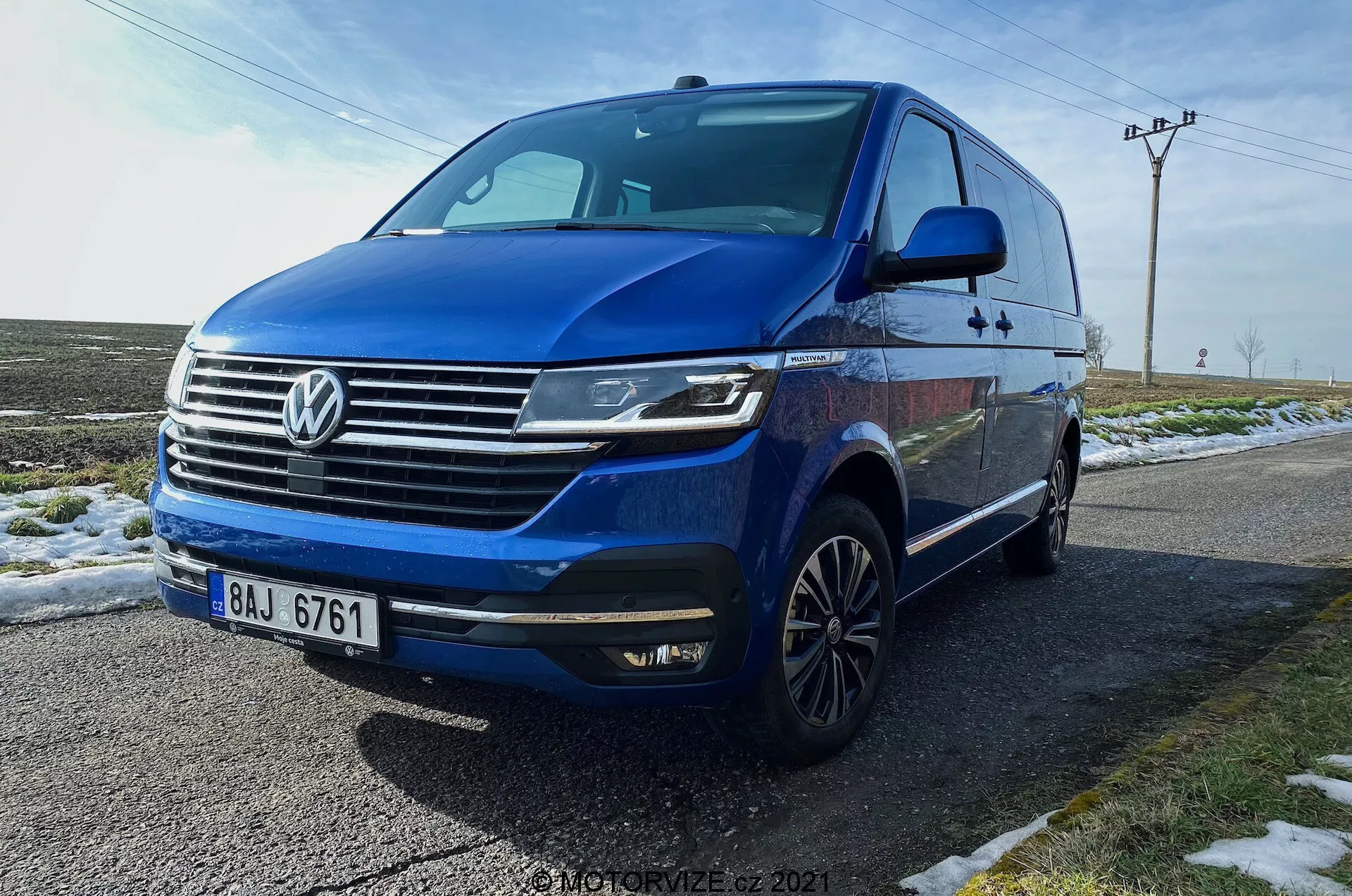 Moder Volkswagen T6.1 Multivan Transporter 2019 je zajet pod nizkim trikotnim kotom spredaj, prikazuje sprednji del avtomobila, masko z izrazitim logotipom Volkswagen, vodoravnimi kromiranimi letvami, kotne LED žaromete z dnevnimi lučmi, sprednji odbijač z vgrajenimi meglenkami in zračniki, dinamično linijo vzdolž strani, večkraka platišča iz lahke zlitine, integrirane smernike na stranskih ogledalih, zatemnjena okna. Vozilo je parkirano na mokri cesti poleg zasnežene trave, z ruralnim ozadjem, ki vključuje električne drogove in odprto nebo.