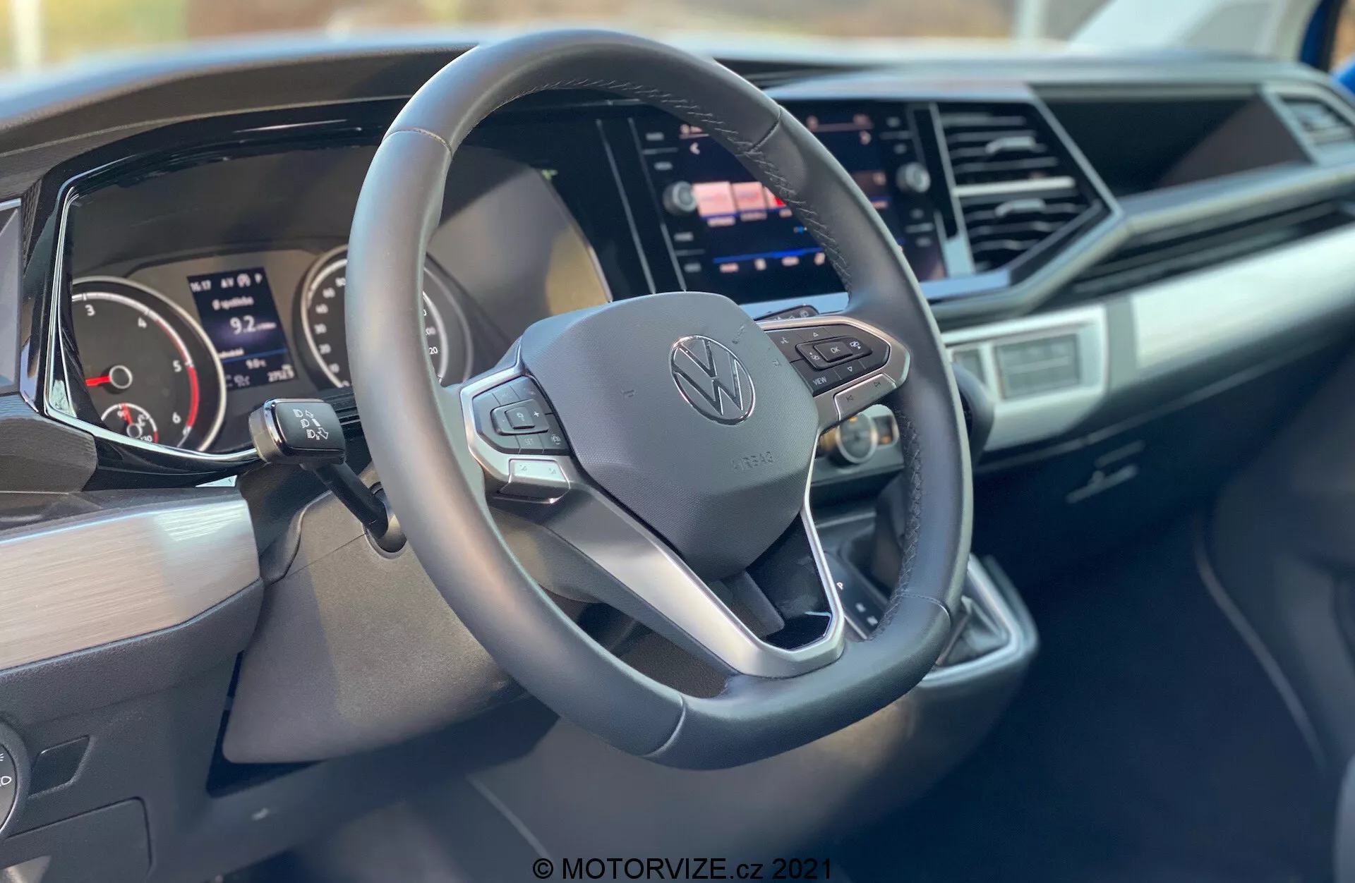 Notranji posnetek avtomobila Volkswagen Nutzfahrzeuge Transporter 2019 (T6-1 Multivan), osredotočen na volan in armaturno ploščo. Posnetek od blizu, posnet z vidika sovoznika, poudarja trikraki, v spodnjem delu ploščati volan z gumbi za upravljanje in osrednjim Volkswagen logom, digitalno instrumentno ploščo, velik osrednji zaslon na dotik za infozabavni sistem, eleganten dizajn armaturne plošče z oglato poudarki in blagovno znamko na sovoznikovi strani armaturne plošče.