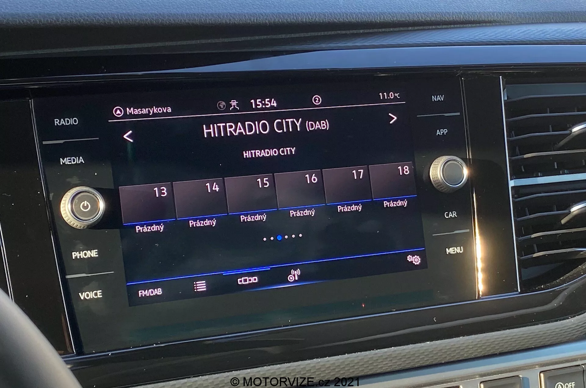 Bližnji sprednji posnetek zaslona infozabavnega sistema v VW-Nutzfahrzeuge Transporter 2019 (T6-1 Multivan). Slika prikazuje izbrano digitalno radijsko postajo 'HITRADIO CITY' z različnimi dotikalnimi možnostmi in funkcijskimi gumbi, kot so 'PHONE', 'VOICE' in 'MENU'. Zaslon prikazuje čas '15:54' in zunanjo temperaturo '11.0°C' z ikonami za uporabniške profile, nastavitvami vozila in močjo signala. Zaslon je vgrajen v osrednji del armaturne plošče, obdan z otipnimi gumbi in kontrolnimi gumbi.