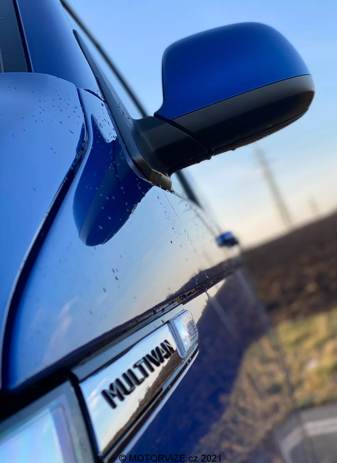 Povečan posnetek strani modrega VW Nutzfahrzeuge Transporter 2019 (T6-1-Multivan), ki vključuje del sprednjih vrat sovoznika, stransko ogledalo in del zadnjih vrat. Poudarek je na oznaki 'MULTIVAN' na avtomobilu, medtem ko je aerodinamično oblikovano stransko ogledalo nekoliko zamegljeno. Ohišje ogledala je enake barve kot barva karoserije, na površini pa so vidne kapljice vode, kar nakazuje na vodoodbojne lastnosti ali nedavni dež.