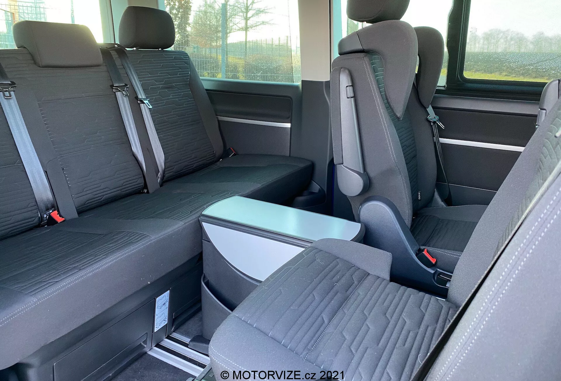 Notranji pogled na VW-Nutzfahrzeuge Transporter 2019 (T6.1 Multivan) s poudarkom na sedežih druge in tretje vrste. Fotografirano od strani na višini oči, slika prikazuje posamezne sedeže z izstopajočimi nasloni za glavo v drugi vrsti in klop za tri potnike v tretji vrsti. Sedeži so dvobarvni, tla pa imajo kovinske tirnice za rekonfiguracijo sedežev. Vidni so zložljiv naslon za roke in sponke varnostnih pasov. Delno sta vidni tudi stransko okno in shranjevanje.