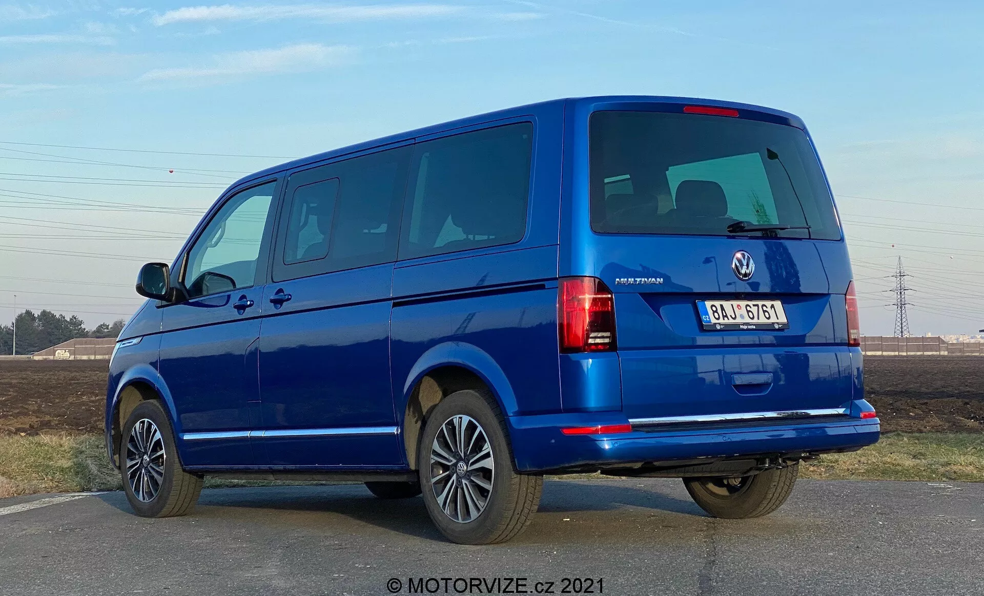 Modri VW-Nutzfahrzeuge Transporter 2019 (T6-1 Multivan) prikazan iz tričetrtinske zadnje perspektive. Slika zajema zadnje luči, zadnja okna in zadnja vrata, ter bočni profil z vrati in okni za potnike. Kombi zaseda velik del okvirja na višini oči, razkrivajoč deljena zadnja vrata in več stranskih oken, kar kaže na potniški model. Platišča iz lahke litine, odbijači v barvi karoserije in stranska ogledala nakazujejo na višjo opremo. Osvetlitev nakazuje na dnevni čas z jasnim nebom, gladkimi sencami pod vozilom.