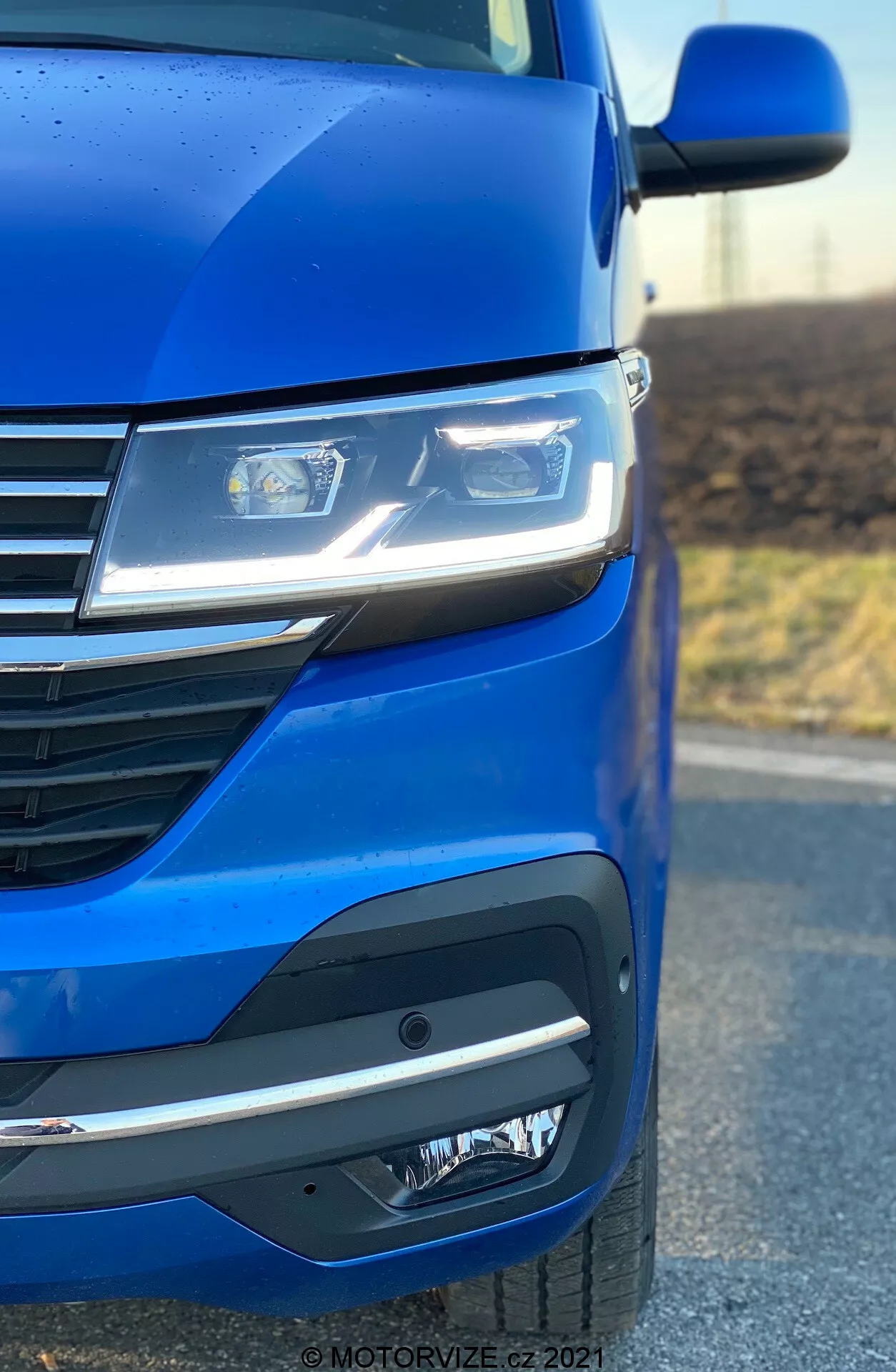 Posnetek od blizu leve sprednje strani vozila VW Nutzfahrzeuge Transporter 2019 (T6.1 Multivan), ki poudarja kovinsko modro barvo, napredne LED žaromete, moderno zasnovo mreže s horizontalnimi črtami in prefinjen sprednji odbijač z meglenkami ali senzorji. Kot fotografije poudarja aerodinamiko in dizajnerske podrobnosti avtomobila.