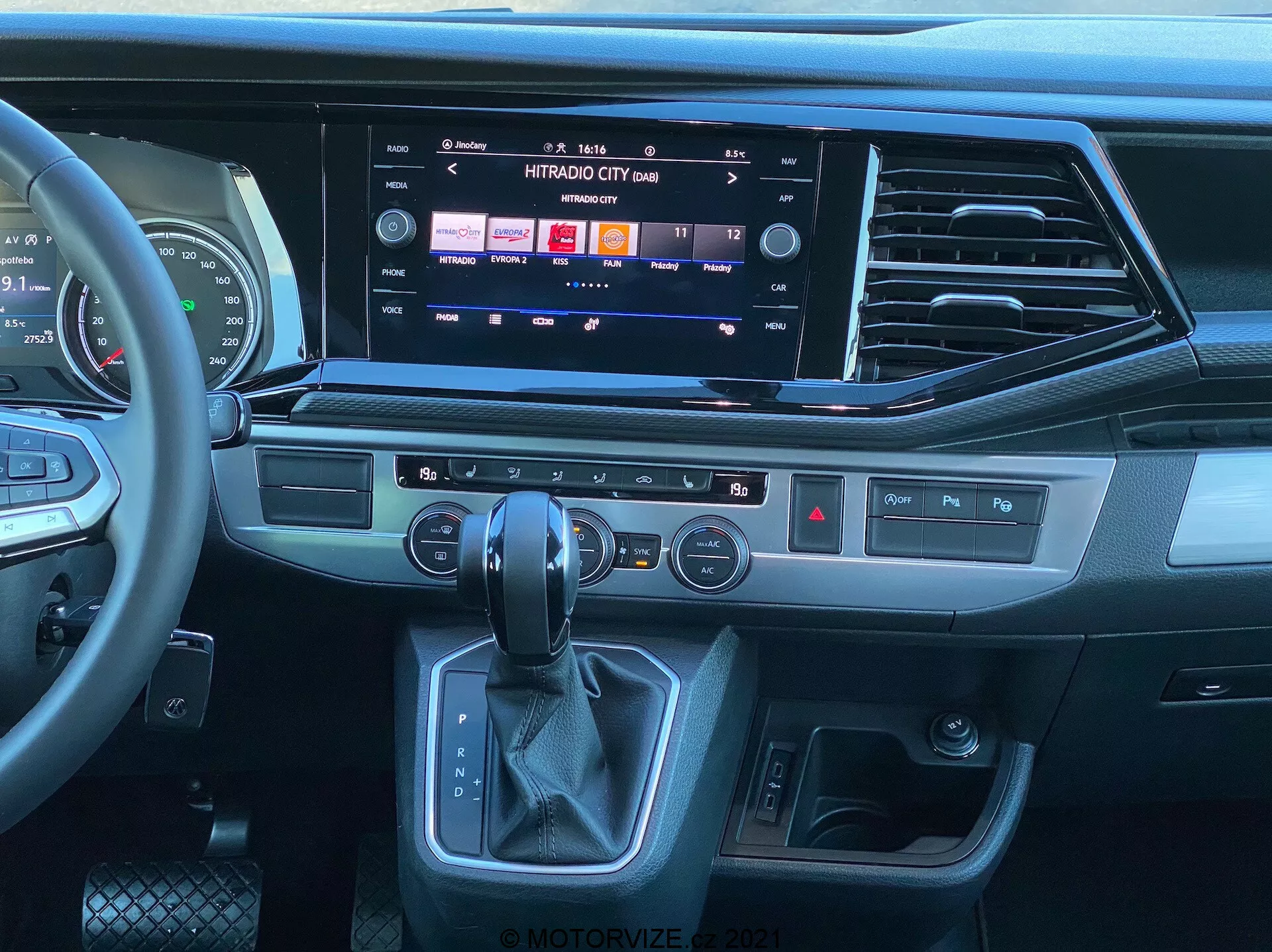 Pogled na notranjost VW Nutzfahrzeuge Transporter 2019 T6.1 Multivan iz perspektive potnikovega sedeža, ki prikazuje sodobno armaturno ploščo in sredinsko konzolo. Večfunkcijski usnjen volan z upravljanjem medijev, velik zaslon na dotik informacijskega sistema, ki prikazuje možnosti medijev in povezljivosti, elegantne zračne odprtine, taktilni gumbi za hitre funkcije, panel za nadzor dvojne klimatske naprave, ergonomski avtomatski menjalnik in gumb za zagon/ustavitev brez ključa. Oblika vključuje kombinacijo temnih plastik, oblog, ki izgledajo kot brušena kovina, in materialov podobnih usnju, kar poudarja sodobni in premium občutek.