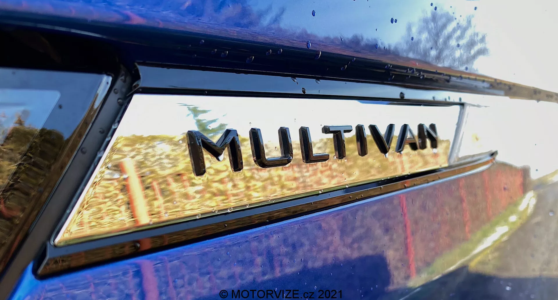Povečava pogled na del VW Nutzfahrzeuge Transporter 2019 (T6-1 Multivan) s poudarkom na znački 'MULTIVAN'. Slikano z nizkega zadnjega kota, nekoliko navzgor, kjer se zadnje stransko okno sreča z robom strehe. Rahli odsevi na steklu okna nakazujejo zunanje okolje. Ta tesno obrezana slika poudarja oznako modela proizvajalca.