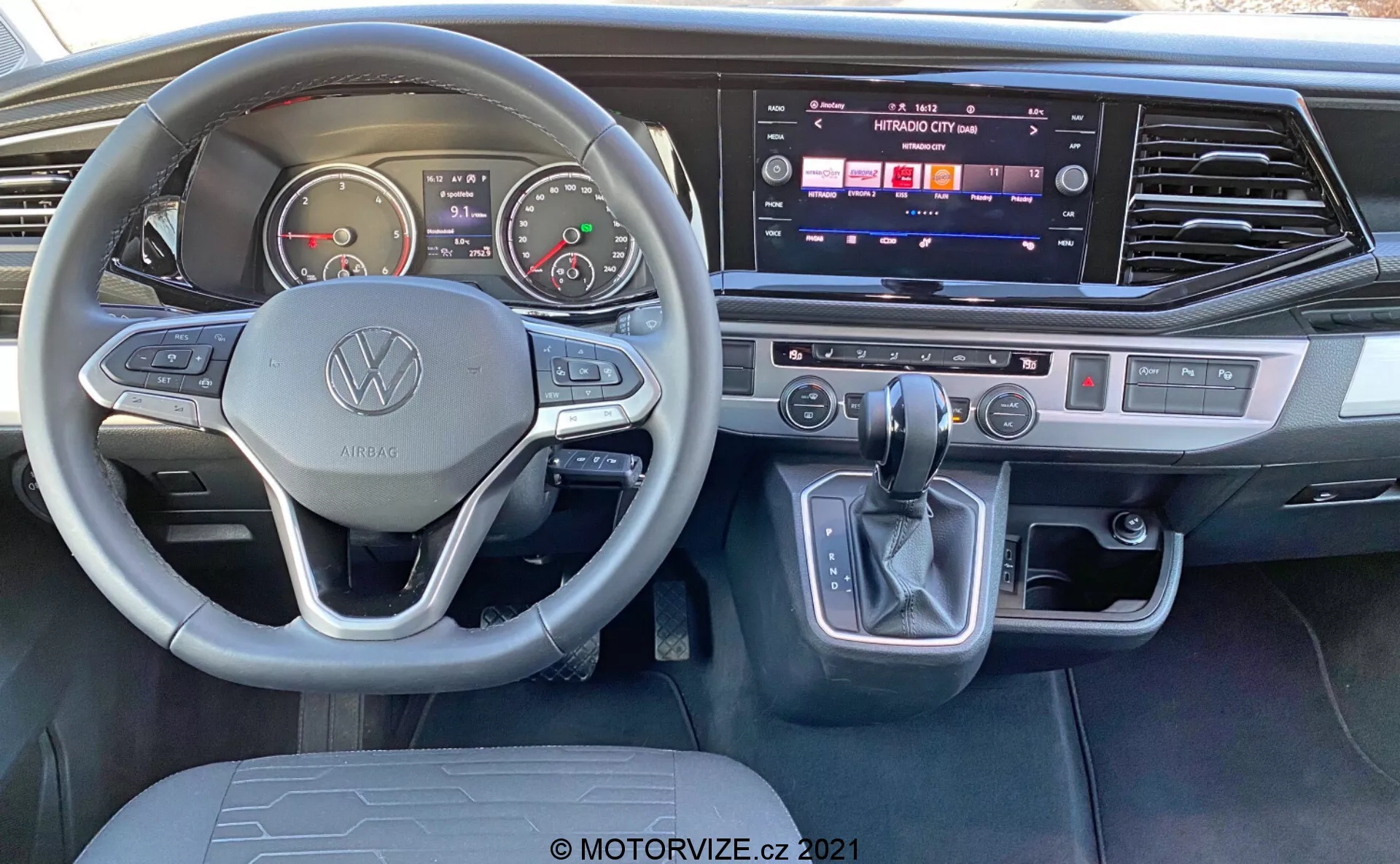 Notranji pogled na VW Nutzfahrzeuge Transporter 2019 T6.1 Multivan z vidika voznikovega sedeža. Vsebuje volan z Volkswagenovim logotipom in večfunkcijskimi gumbi, analogne merilnike, digitalni zaslon, sredinsko konzolo z aktivnim zaslonom na dotik za informacijsko-razvedrilni sistem, dvopodročno klimatsko napravo, prestavno ročico z elektronskim parkirnim gumbom in moderne brušene kovinske poudarke.