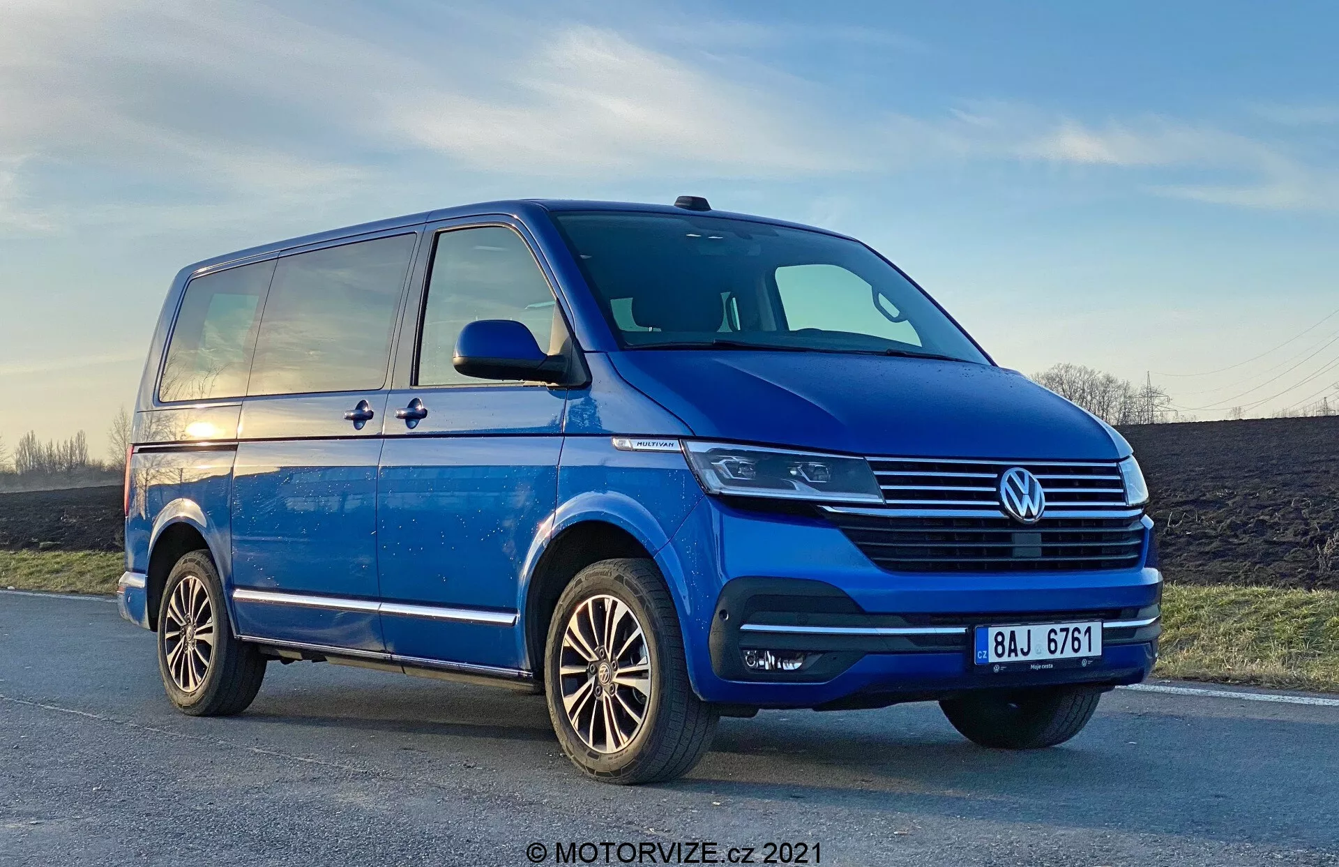 Nizek kotni pogled na modri Volkswagen T6-1 Multivan 2019, ki prikazuje prednji levi kvadrant iz tričetrtinskega vidika. Kombi je parkiran na rahlo nagnjeni cesti, z vidnimi kromiranimi detajli na rešetki, centralnim VW emblemom in sodobnimi žarometi, ki lahko uporabljajo LED tehnologijo. Meglenke so integrirane v odbijač, ki ima tudi zračne dovode. Bočni profil razkriva drsna vrata, linijo karakterja, ki se razteza od sprednjega blatnika do zadka, ter robustna stranska ogledala s smerniki. Vidna so tudi alu platišča z večkrakim dizajnom in dobro vzdrževanimi tekalnimi plastmi. Nebo v ozadju kaže nekaj sončnega zahoda.