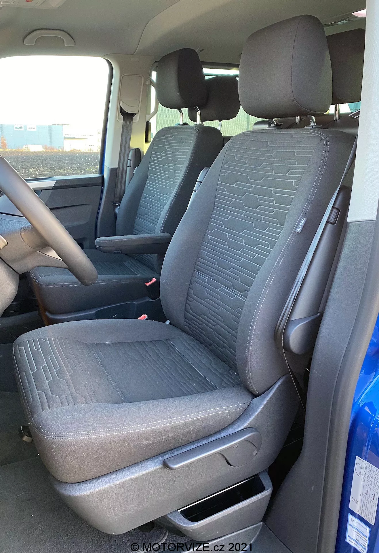 Notranjost VW Nutzfahrzeuge Transporter 2019 T6.1 Multivan, ki prikazuje sprednji sovoznikov sedež in del voznikovega sedeža iz zunanje perspektive na sovoznikovi strani. Posnetek, narejen iz rahlo nižjega kota, poudarja teksturirano tkanino z geometrijskimi vzorci na sedežih, ročico za ročno nastavitev, varnostne pasove in del sredinske konzole ter volana. Naravna dnevna svetloba osvetljuje notranjost vozila.