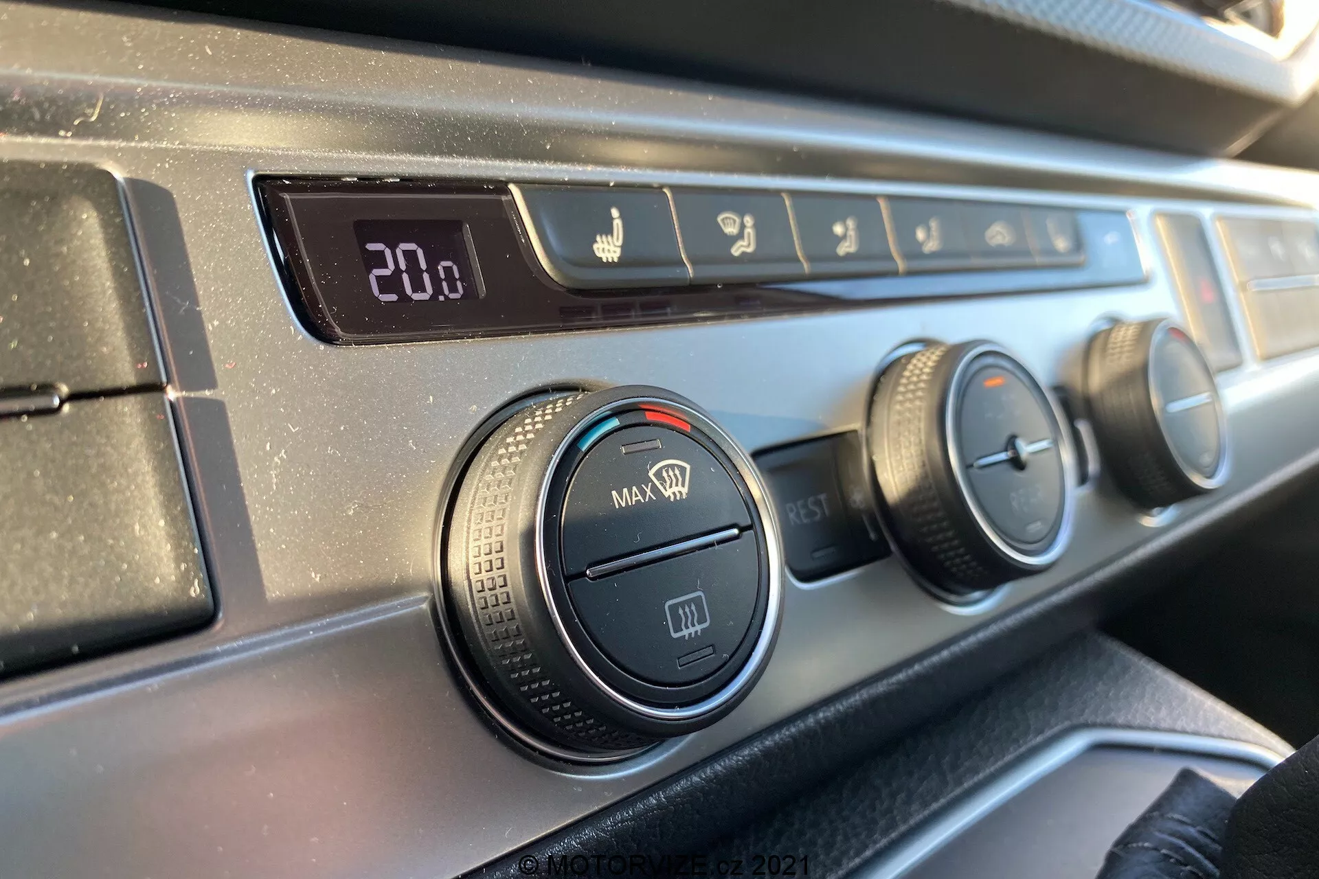 Pogled od blizu na nadzorno ploščo za klimatsko napravo na armaturni plošči VW Transporter T6.1 Multivan 2019 z vidika potnika na sprednjem sedežu, z digitalnim prikazom temperature nastavljene na 20,0 stopinj Celzija, krmilniki za smer zračnega toka, tremi vrtljivimi gumbi za nastavitve klimatske naprave in ventilatorja ter gumbom za recirkulacijo, vse s teksturiranim črnim plastičnim robom in gladkim temnim zaključkom armaturne plošče.