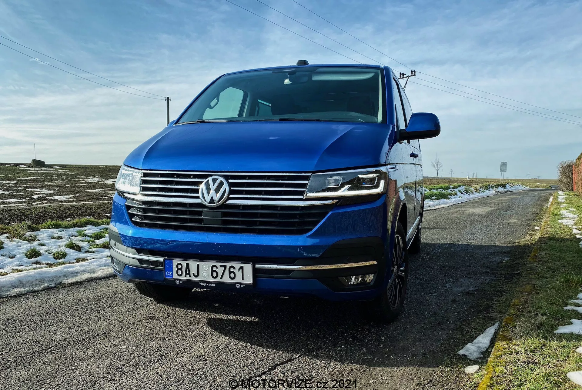 Nizek sprednji tristranski pogled na Volkswagen T6.1 Multivan 2019, ki prikazuje sprednjo rešetko s horizontalnimi črtami, značilno VW oznako na sredini rešetke, žaromete verjetno z LED dnevnimi lučmi in bi-ksenon ali LED lučmi, ter sprednji odbijač z meglenkami in zračnimi odprtinami. Slika prikazuje del leve strani, vključno z voznikovimi vrati in blatnikom, s kapuco in vetrobranskim steklom, ki je vidna. Avto je temno moder in parkiran na podeželski cesti s snegom na tleh in s čisto modrim nebom nad njim.