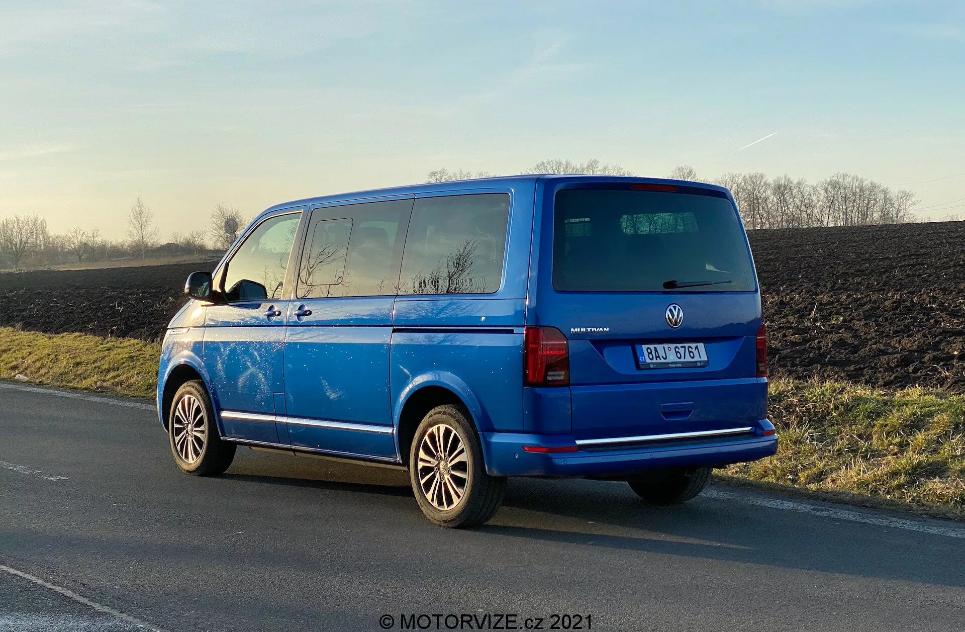 Svetlo modri Volkswagen Transporter 2019 (T6-1 Multivan) parkiran ob cestišču, zadnji tridelni pogled prikazuje zadnje luči, zadnje okno, zadnja vrata, proizvajalčev znak in podrobnosti modela; vidna aluminijasta platišča, zunanja stranska ogledala, linije grelnika zadnjega okna, zatemnjena druga vrsta oken, strešna antena; parkiran na naravni dnevni svetlobi z odsevi, ki poudarjajo polirano površino, podeželsko ozadje.