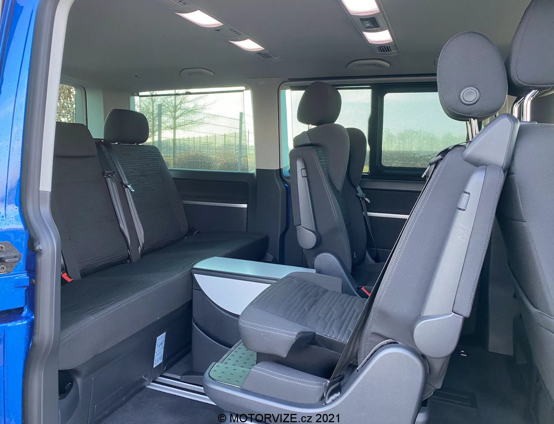 Notranjost VW Transporter T6.1 Multivan 2019, ki prikazuje drugi in tretji sedežni vrsti iz zadnje potniške perspektive, s sedežno konfiguracijo 2-3-2 in temno tkaninsko oblazinjenjem. Delni pogled na zunanjost skozi odprta vrata, vzglavniki na vseh sedežih, dvignjeni sedeži v tretji vrsti za boljšo vidljivost.