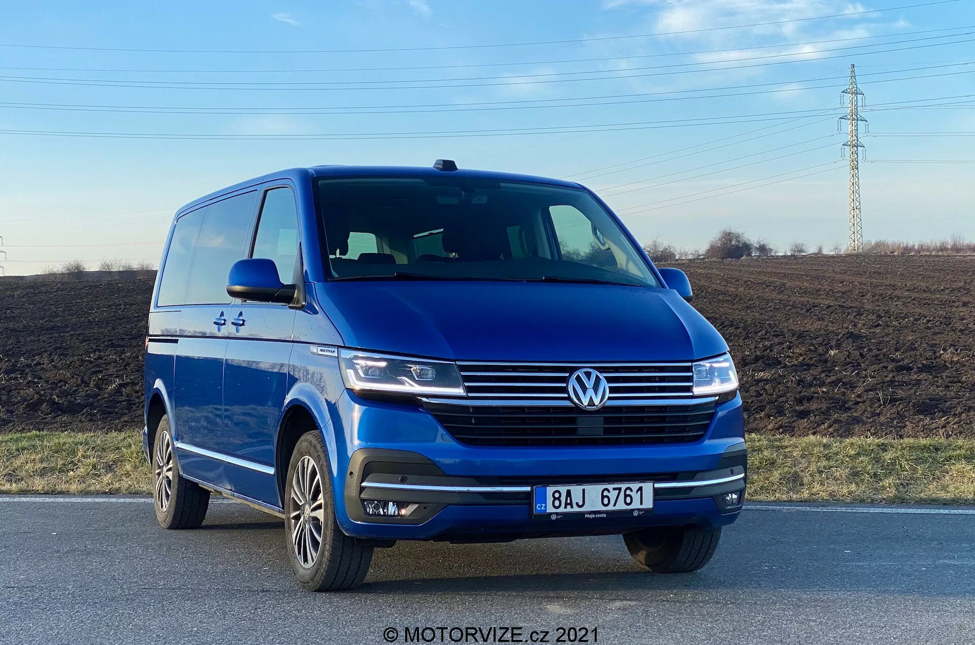 Slika prikazuje moder Volkswagen VW-Nutzfahrzeuge Transporter 2019 (T6-1 Multivan), posnet iz nizkega sprednjega kota, ki prikazuje sprednjo levo četrtino z jasnim pogledom na sprednji del in voznikov profil. Vozilo se nahaja na cesti, izpostavljajoč elemente, kot so izraziti znak Volkswagen, elegantni žarometi, oblikovane linije na pokrovu motorja, integrirane meglenke na odbijaču in večkraka aluminijasta platišča. Fotografija, posneta podnevi, poudarja zunanji dizajn vozila in tehnologije za pomoč vozniku, kot so parkirni senzorji.