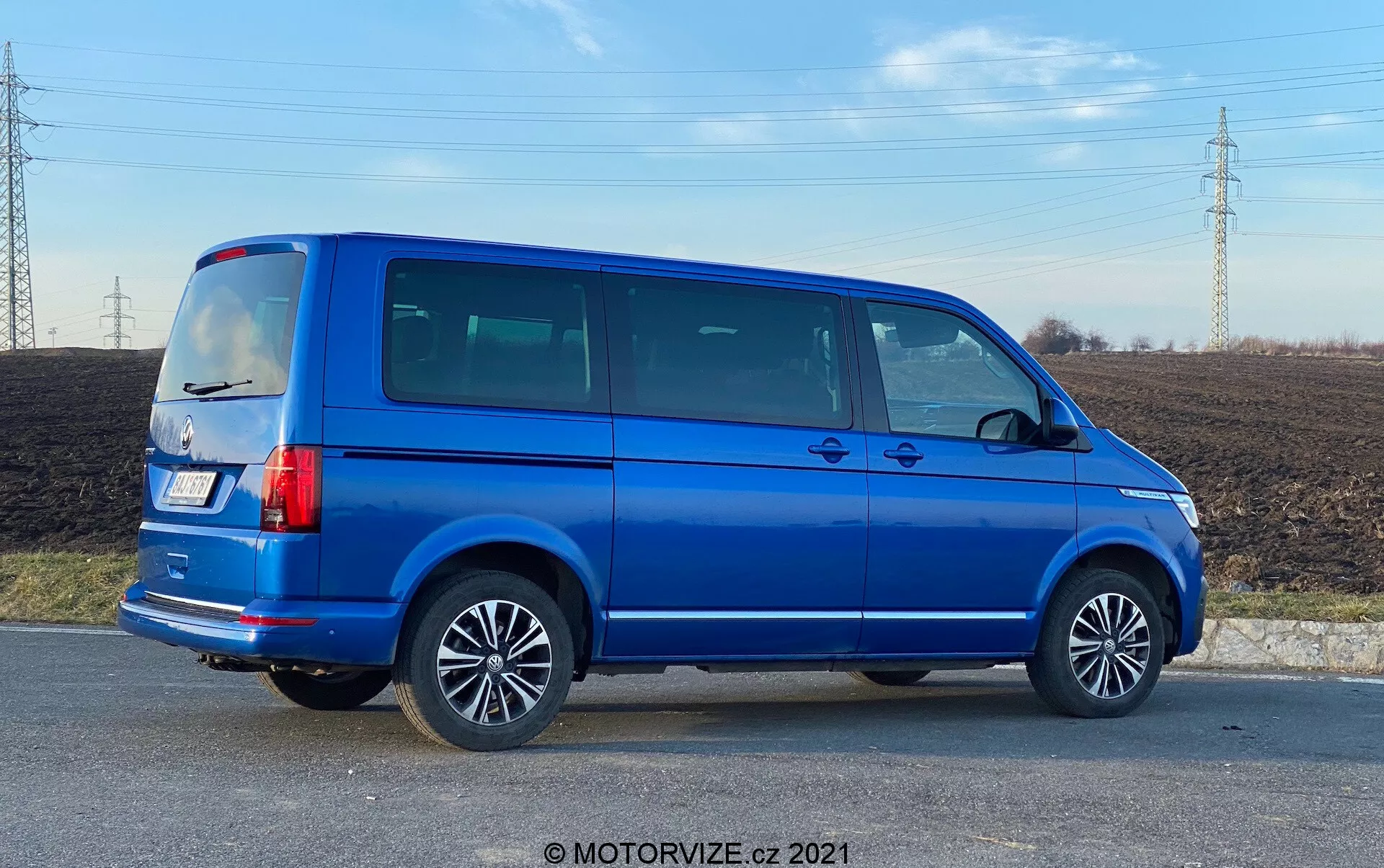 Popoln stranski profil modrega VW Nutzfahrzeuge Transporter iz leta 2019 (T6.1 Multivan) z aluminijastimi platišči, vidnimi linijami vrat, zatemnjenimi stranskimi okni, kvadratno silhueto, navpičnimi zadnjimi lučmi, anteno na strehi in svetlo modro kovinsko barvo. Slika je posneta pod kotom, kar nudi perspektivo tako na stranski kot zadnji del vozila, postavljena v podeželsko območje s poljem in električnimi vodi v ozadju, podnevi.