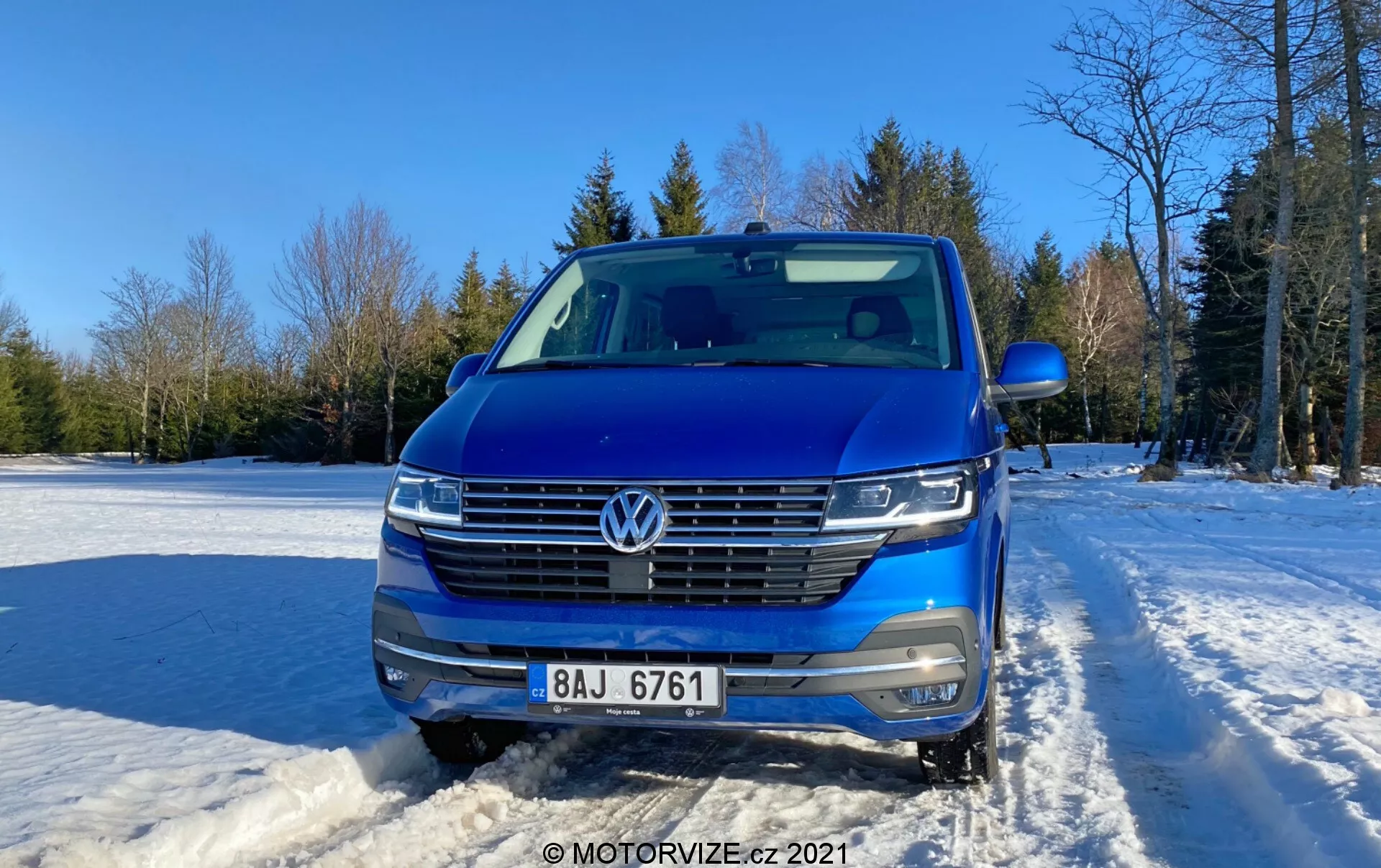 Frontalni pogled na modri Volkswagen Transporter 2019 (T6-1 Multivan), ki prikazuje masko, žaromete in pokrov motorja. Vozilo je postavljeno na snežni podlagi na prostem z jasno nebo in drevesi v ozadju. Značilnosti avtomobila vključujejo znak VW, vodoravno masko s kromiranimi obrobami, LED žaromete, sprednjo kamero, parkirne senzorje, srebrne strešne letve in delno vidno stransko ogledalo. Sodobno oblikovanje z aerodinamično prednjo fasado, fotografirano podnevi z mehko sončno svetlobo z desne strani.