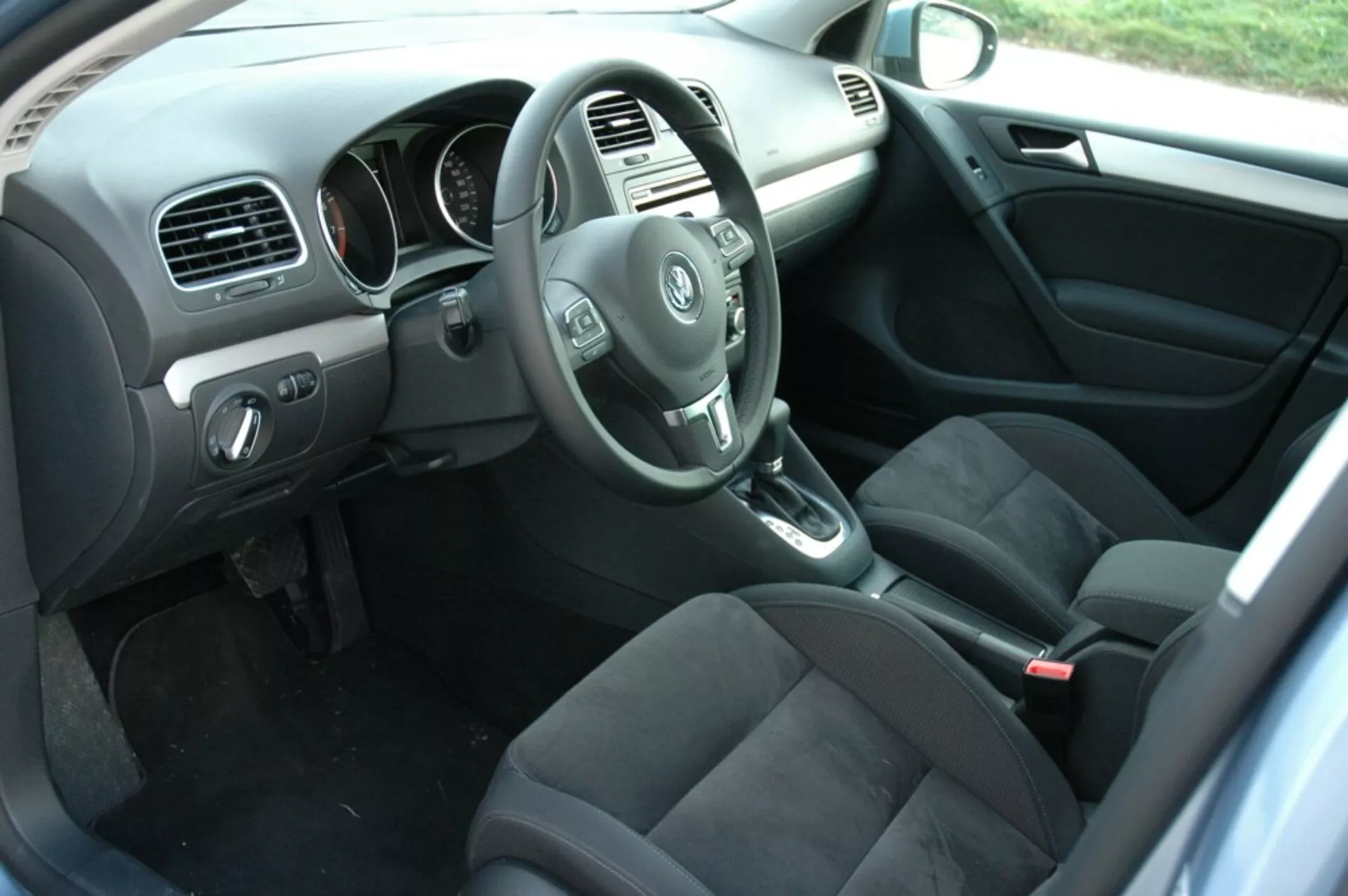 Notranjost VW Golfa 2008, ki prikazuje stran voznika iz perspektive zadnjega potnika, z volanom, armaturno ploščo, sredinsko konzolo, ročico avtomatskega menjalnika, sedeži iz temne tkanine, kontrolami na vratih voznika, preprogo z blazinami in naravno osvetlitvijo