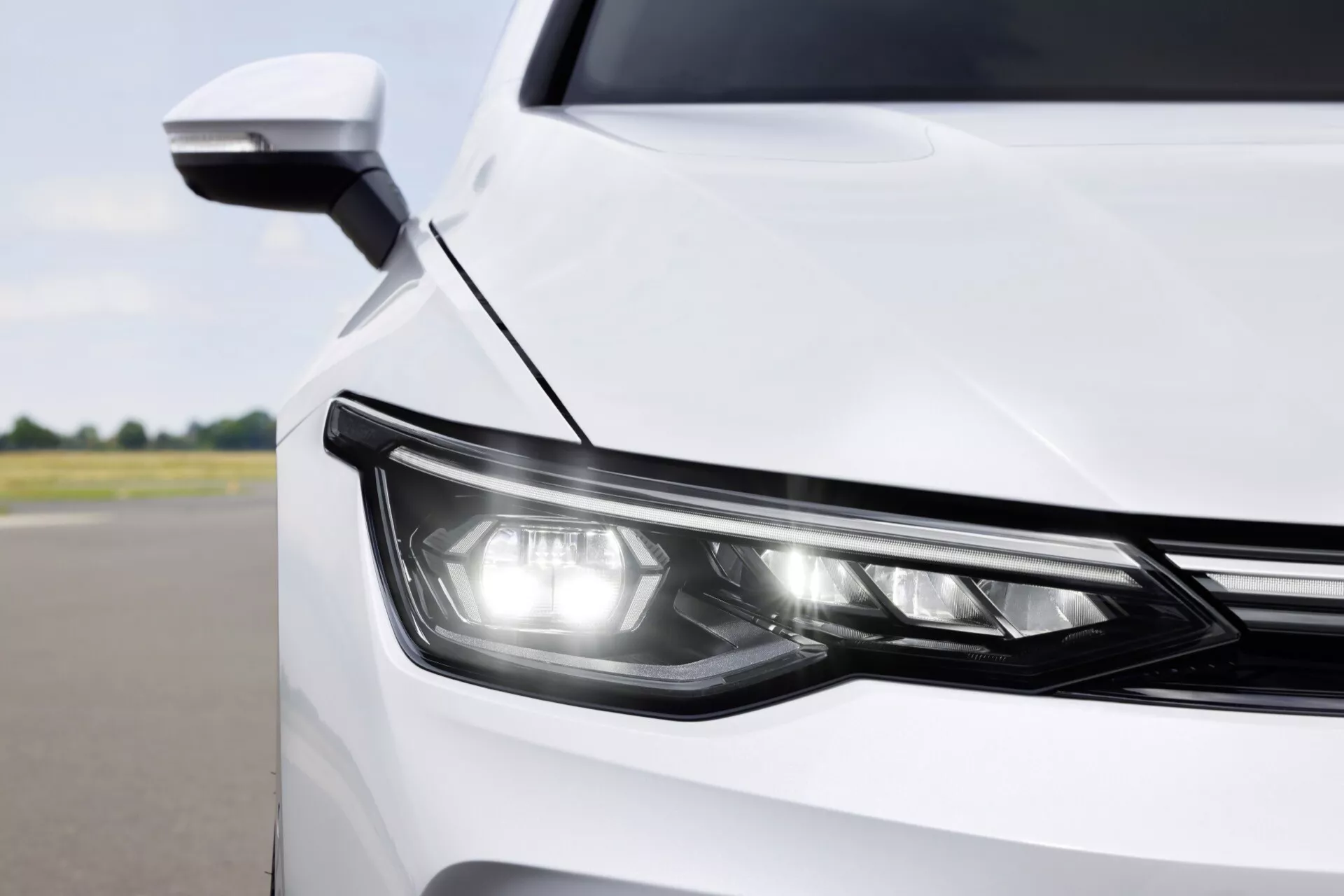 Posnetek sprednjega kota bele VW Golf VIII facelift 2024, ki prikazuje sodobno oblikovanje in tehnologijo LED žarometa in aerodinamičnega stranskega ogledala z integriranim smernikom, iz delnega pogleda pod rahlo povišanim kotom