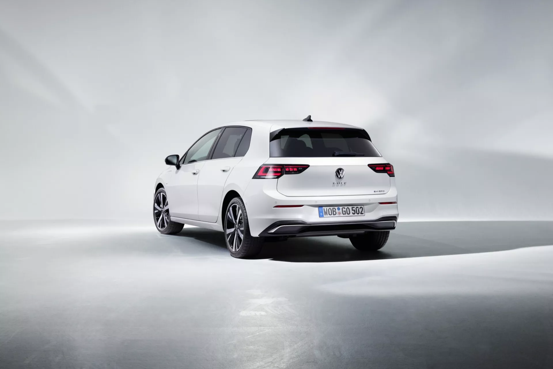 Volkswagen Golf VIII facelift 2024 bele barve, ki prikazuje LED zadnje luči, športni zadnji odbijač z integrirano izpušno konico, obliko hatchback, kakovostna lita platišča in potencialno tehnologijo pomoči pri parkiranju, vse zajeto v dinamičnem treh četrt pogledu od zadaj z mehko osvetlitvijo, ki poudarja oblikovne krivulje na enostavnem ozadju.