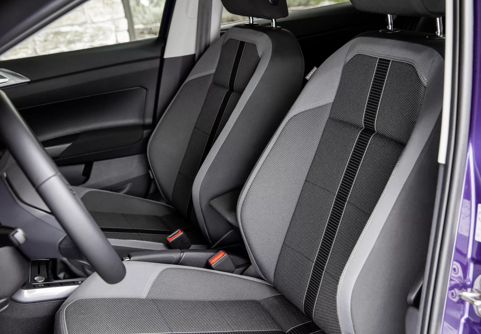 Notranjost VW Polo 2021 (VI-facelift) iz perspektive odprtih sprednjih vrat kaže sprednje in zadnje sedeže. Slika poudarja konfiguracijo sedeža, oblikovanje tkanine, šive in podrobnosti konture. Vidni so nastavljivi vzglavniki, varnostni pasovi in delno voznikov stranski plošček vrat, poudarjajoč udobje in notranjo obliko.