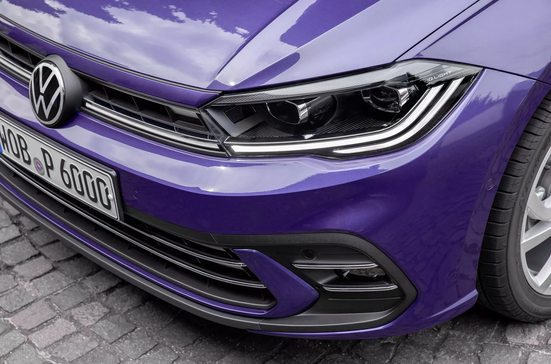 Od blizu, poševen sprednji pogled na modri Volkswagen Polo 2021 VI-facelift, prikaže sodobno zasnovo žarometov z integriranimi LED dnevnimi lučmi, značilen logotip Volkswagen na horizontalni mreži in športni odbijač z rešetkami za dovod zraka in dizajnom s satovjem. Podrobnosti poudarjajo elegantne linije pokrova motorja in bleščečo lakirano površino, kar nakazuje napredno osvetlitveno tehnologijo in potencialne sisteme za pomoč vozniku.