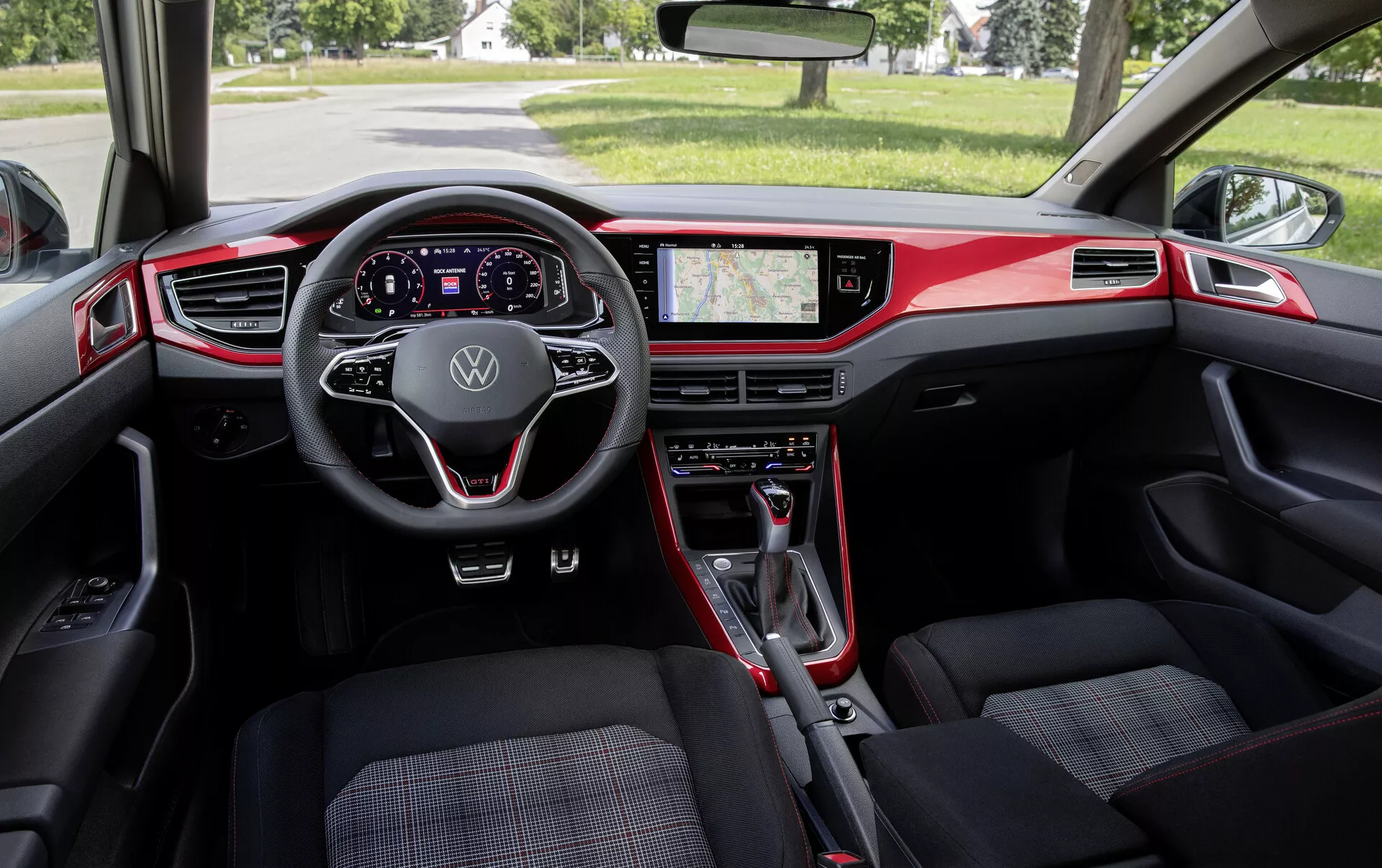 Notranji pogled na VW Polo 2021 (VI-facelift) z vidika zadnjega sedeža. Posnetek iz širokega kota zajema armaturno ploščo, sprednja sedeža in vzvratno ogledalo. Značilnosti vključujejo sodoben večfunkcijski volan z logotipom Volkswagen, popolnoma digitalni instrumentni zaslon, velik zaslon na dotik za informacijsko-razvedrilni sistem z GPS navigacijo in fizične kontrole klimatske naprave. Notranjost, ki je večinoma črna z rdečimi poudarki, poudarja športni dizajn. Prednja sedeža sta oblazinjena z vzorčasto tkanino, prestavna ročica pa kaže na samodejni menjalnik. Notranjost združuje funkcionalnost, sodoben dizajn in napredno tehnologijo.