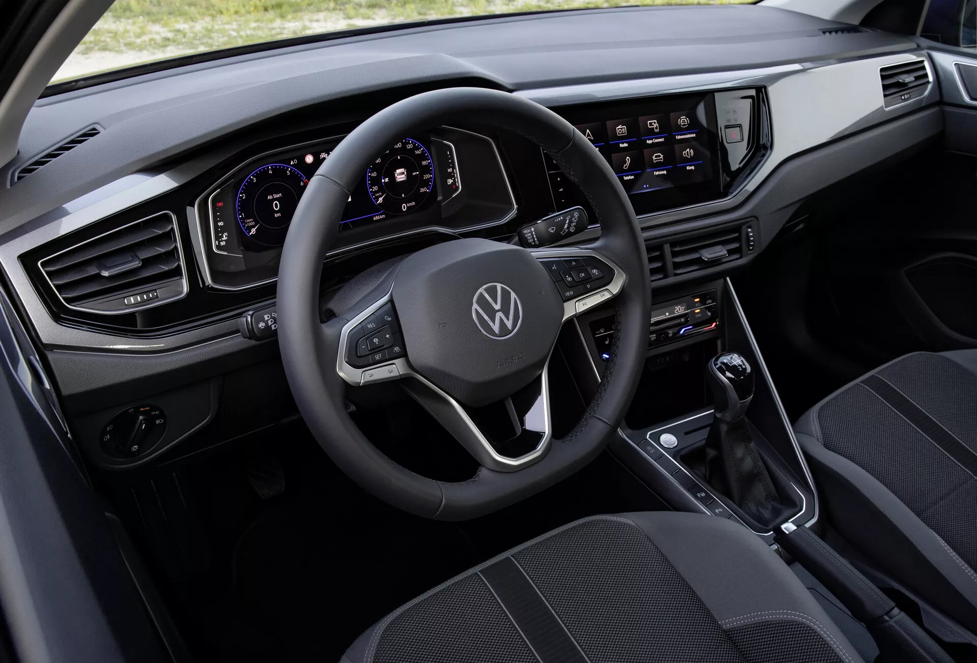 Pogled z zgornje strani notranjosti VW Polo 2021 (VI-facelift), ki prikazuje večnamenski usnjeni volan z ravnim spodnjim dizajnom, okrašen z logotipom Volkswagen, popolnoma digitalni in prilagodljivi instrumentni panel, velik zaslon na dotik z informacijsko-razvedrilnim sistemom, sodoben horizontalen dizajn armaturne plošče z elegantnimi, kotnimi zračniki, minimalistično sredinsko konzolo z ročico za samodejni menjalnik ter visokokakovostne sedeže iz tkanine s vzorčastim dizajnom.