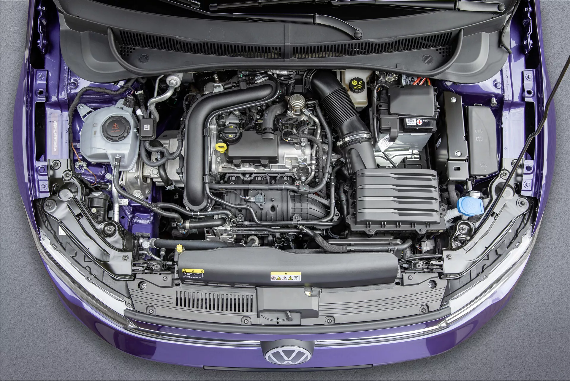 Pogled od zgoraj na Volkswagen Polo 2021 (VI-Facelift) z dvignjenim pokrovom, ki razkriva prečno nameščen motor in različne komponente v motornem prostoru. Slika prikazuje plastični pokrov motorja, baterijo, sesalni sistem zraka, rezervoar za hladilno tekočino z modrim pokrovčkom, različne posode za tekočine, komponente motorja z kovinskimi in gumijastimi cevmi ter varovalko. Barva avtomobila je vijolična in v standardnem tovarniškem stanju. Idealno za kategorije, ki se nanašajo na avtomobilske motorje, vzdrževanje vozil, avtomobilski inženiring in proizvodnjo avtomobilov.