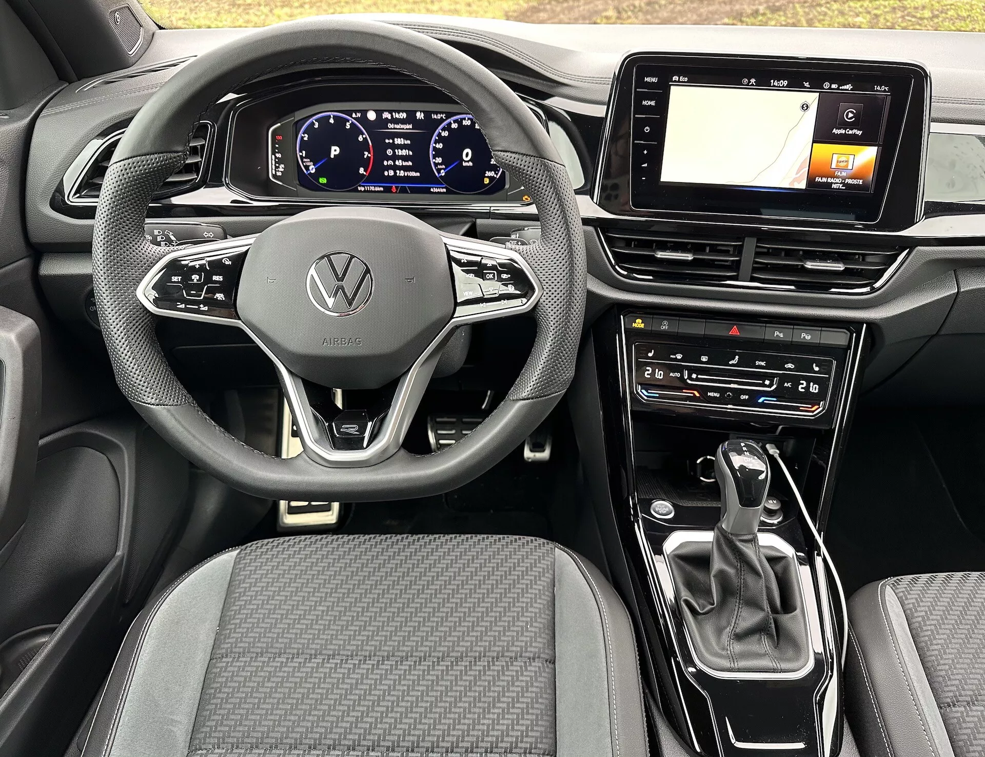 Notranji pogled na VW T-Roc Cabriolet 2022 s sodobnim trorokim volanom z emblemo Volkswagen, digitalnim instrumentnim zaslonom, zaslonom za informacijsko-zabavni sistem z GPS, izbirnikom prestav avtomatskega menjalnika in nastavitvami klimatske naprave, ki poudarjajo napredno tehnologijo in elegantno obliko avtomobila