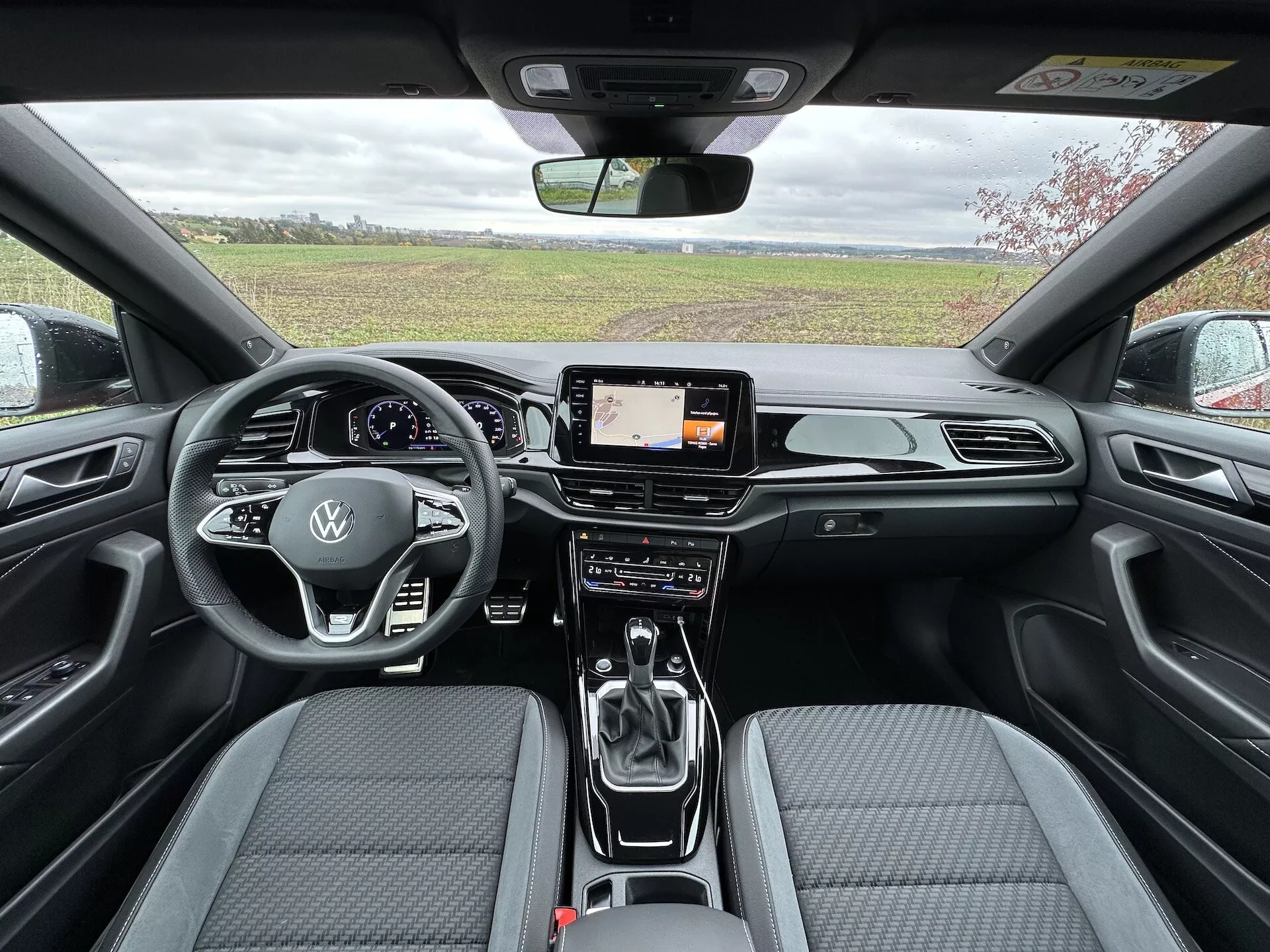 Notranji pogled VW T-Roc Cabriolet 2022 od zadaj prema spredaj prikazuje aerodinamično armaturno ploščo s kromiranimi poudarki, digitalni zaslon zabavnega sistema, napredni dvoconski sistem klimatizacije, večfunkcijski volan oblečen v usnje z logotipom Volkswagen, avtomatsko prestavno ročico, in sodobne sedeže iz dvobarvne tkanine z vzorcem šivov.