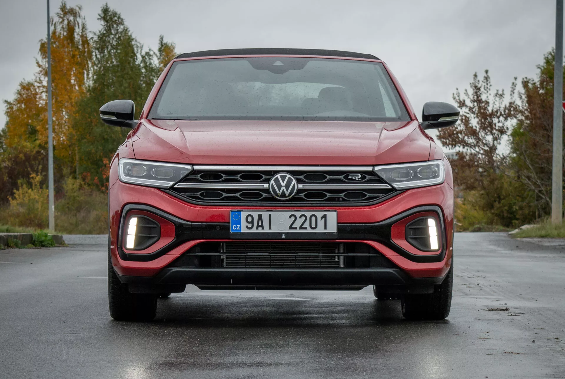 Sprednji pogled na VW T-Roc Cabriolet 2022, ki poudarja elegantno masko z logotipom Volkswagen, LED žaromete, spodnji zračnik z rdečim poudarkom za športen videz in aerodinamično zasnovo na mokrem cestišču