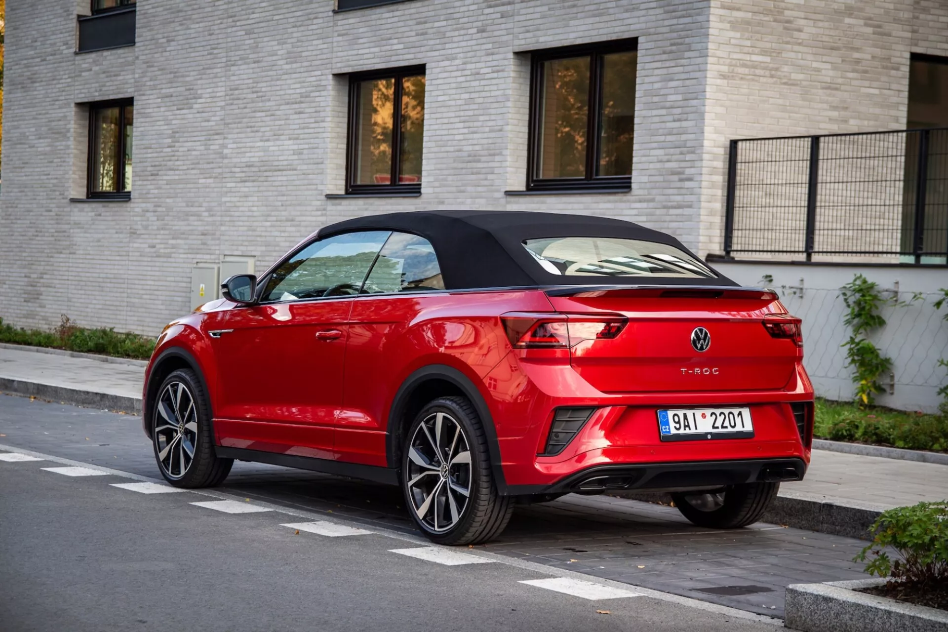 Zadnji tričetrtinski pogled na rdeč Volkswagen T-Roc Cabriolet, parkiran na ulici, ki poudarja njegovo črno zložljivo streho, elegantne LED zadnje luči, zadnji spojler, dvojne izpušne cevi in integrirane parkirne senzorje na ozadju svetlo obarvane opečnate stavbe.