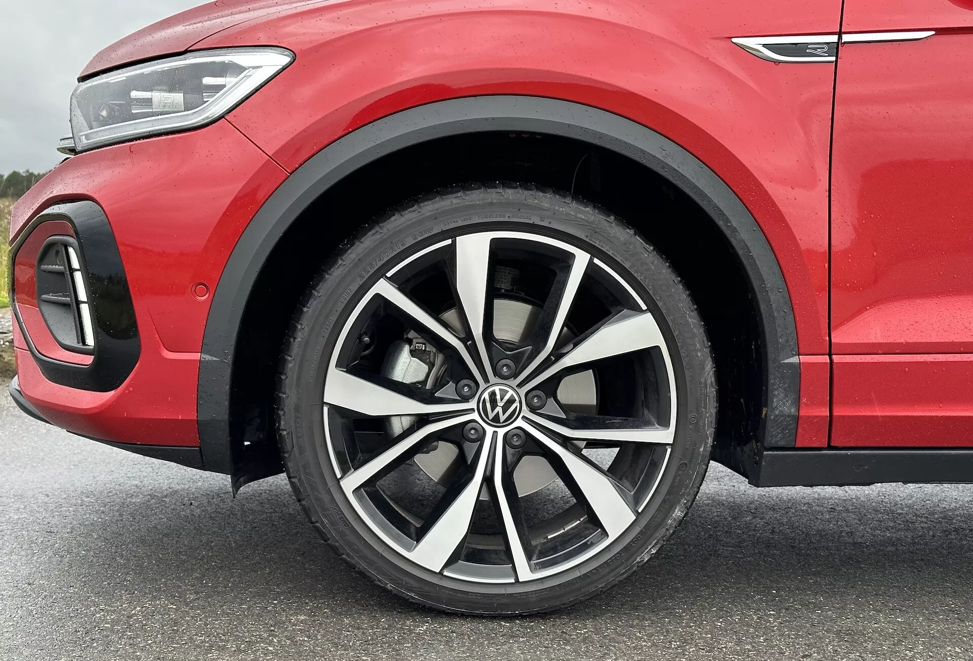 Podroben pogled na VW T-Roc 2022 s poudarkom na večkraki zlitinski kolesi z dvobarvno obdelavo, nizkoprofilno pnevmatiko in delno sprednjega blatnika, ki prikazuje športno rdečo karoserijo in podrobnosti LED osvetlitve