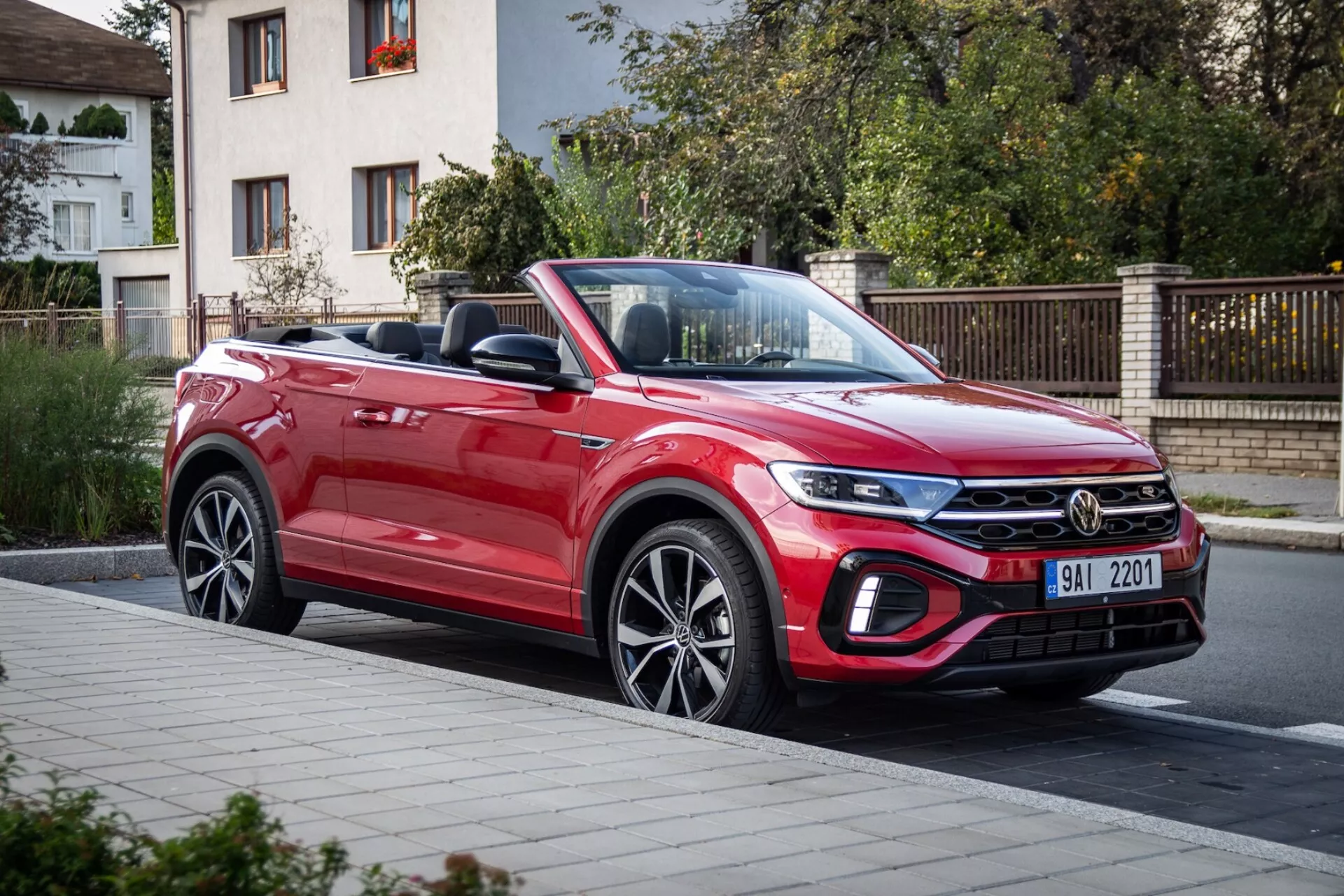 Rdeči kabriolet VW T-Roc 2022, ki predstavlja svojo dinamično zasnovo z dolgim padajočim pokrovom motorja, moderno sprednjo rešetko, elegantnimi žarometi in aerodinamičnim profilom, ujetim iz treh četrtin sprednjega pogleda na cesti
