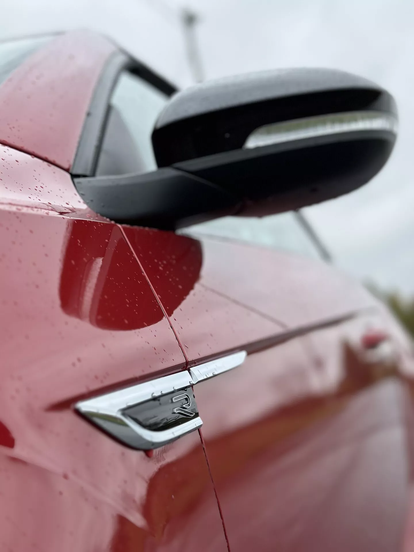 Podrobn pogled s strani na rdeč VW T-Roc Cabriolet 2022, ki poudarja zadnji del z detajlnim pogledom na črno stransko ogledalo, kljuko vrat z brezključnim vstopom in oblikovalske elemente avtomobila, vključno z značko, pod rahlo navzgor nagnjenim kotom z vodnimi kapljicami na površini, ki kažejo na nedavni dež ali pranje, proti zamegljenemu ozadju