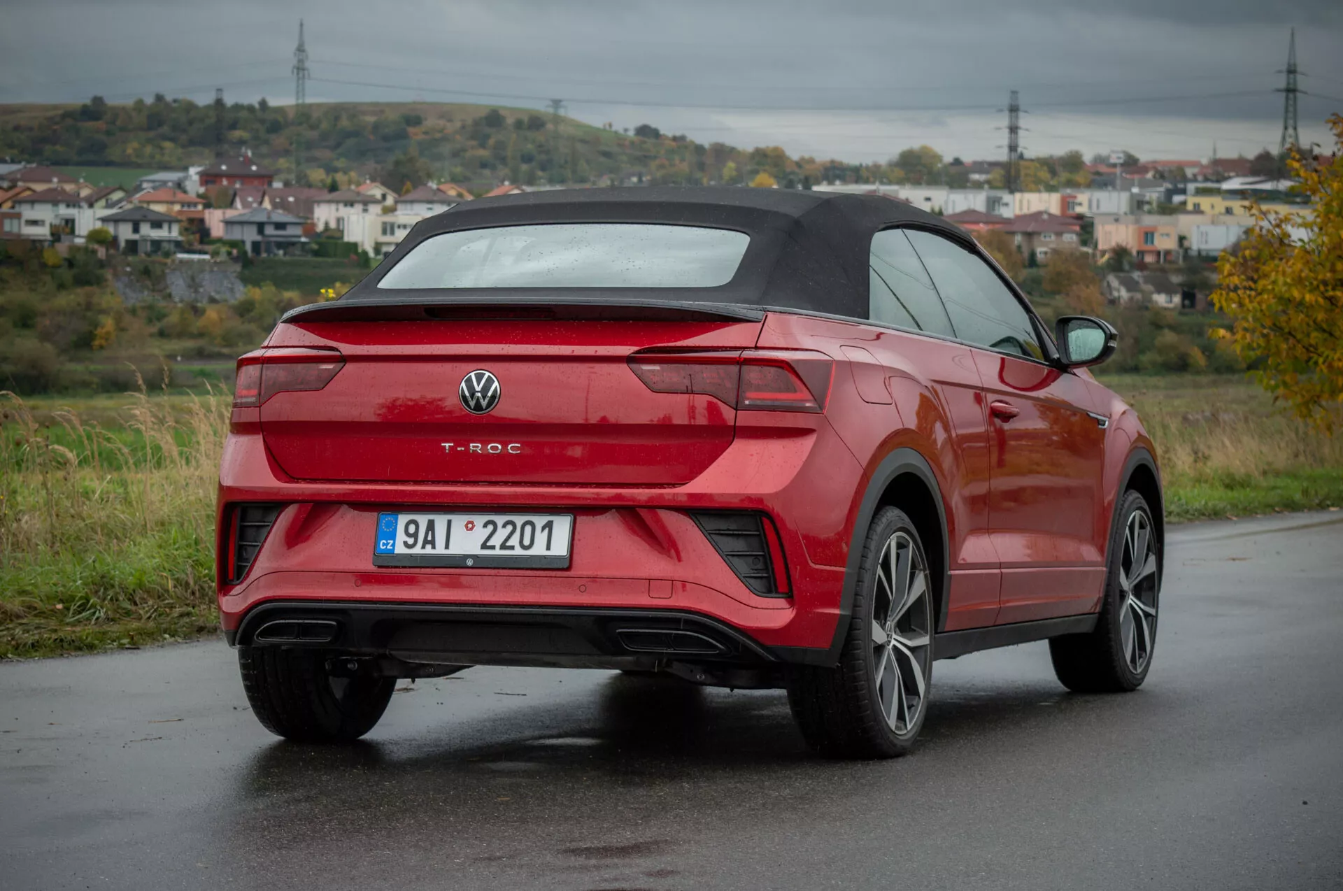 Rdeč Volkswagen T-Roc Cabriolet 2022 v tričetrtinskem zadnjem pogledu na mokri cesti, ki prikazuje zadnji dizajn, stranski profil, oblikovanje koles, LED zadnje luči, zadnji odbijač z integriranimi senzorji, dvojne izpušne cevi, večkraka lita kolesa in zložljivo streho