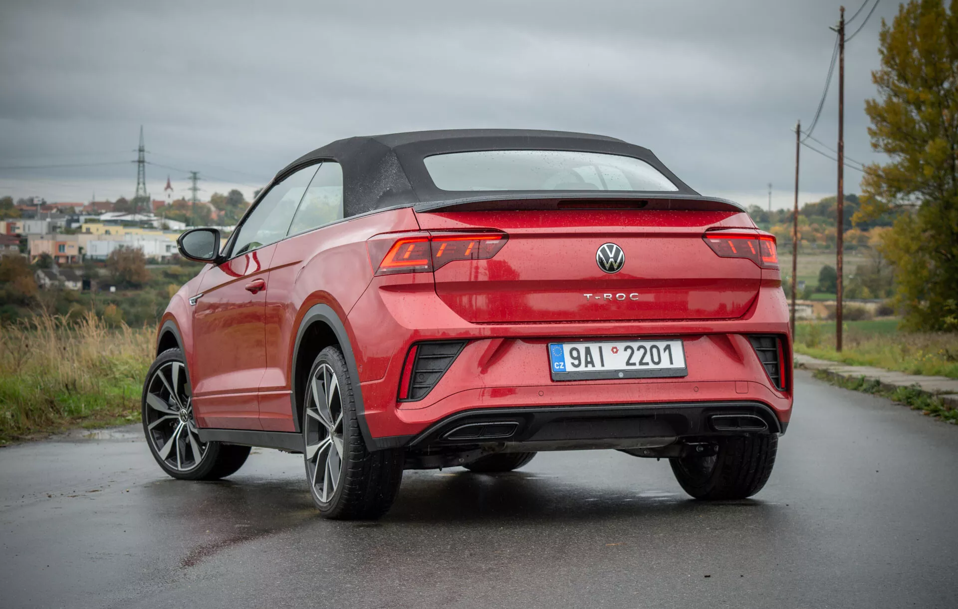 Zadnji tričetrtinski pogled na VW T-Roc Cabriolet 2022, parkiran na mokri cesti, ki prikazuje njegov sodoben kabrioletski dizajn, ostre linije karoserije, športna lita kolesa, elegantne zadnje luči in dvojni izpuh, v ozadju pa polpodeželsko okolje z električnimi stebri in bujnim rastlinjem.