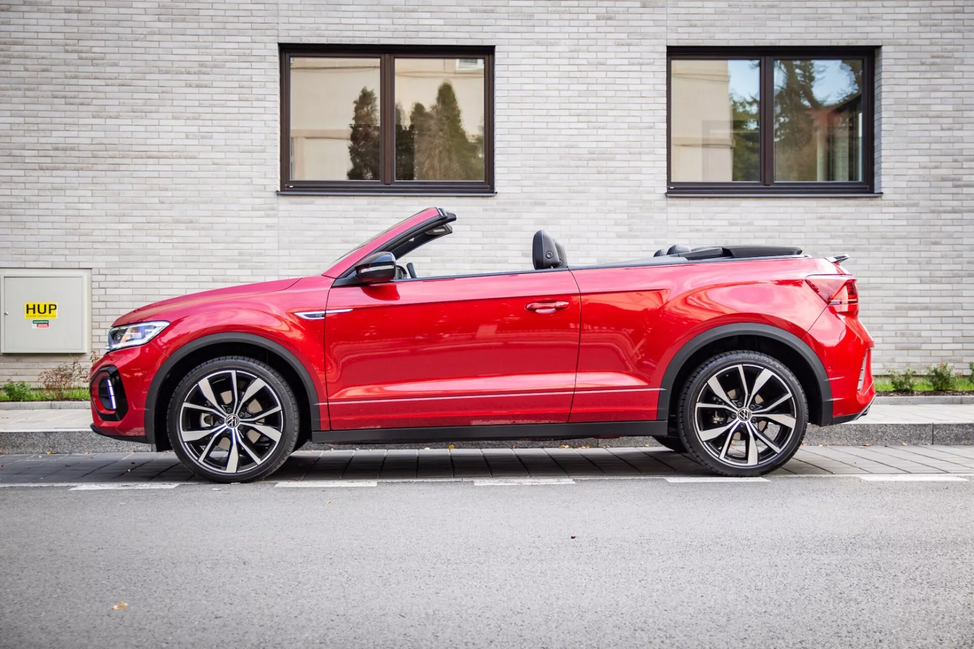 Rdeči kabriolet Volkswagen T-Roc 2022 parkiran vzporedno s sivo opečnato steno z znakom HUP, prikazuje stranski profil z rahlim sprednjim kotom, spuščeno streho kabrioleta, LED luči in športna lita platišča