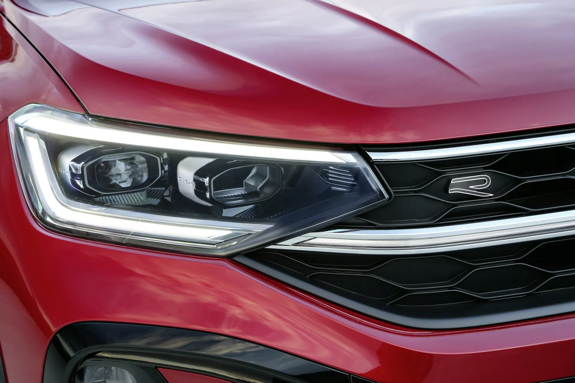 Približni pogled na rdeč VW Taigo 2021, ki poudarja levi žaromet, sprednji odbijač in del rešetke s kromiranimi poudarki, prikazuje LED osvetlitev, sodobno oblikovanje in športni, luksuzni videz